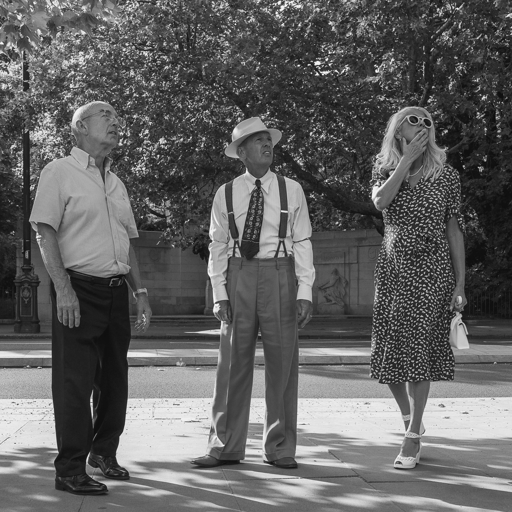 Three people standing outdoors under trees, dressed in vintage or formal attire, looking in different directions.