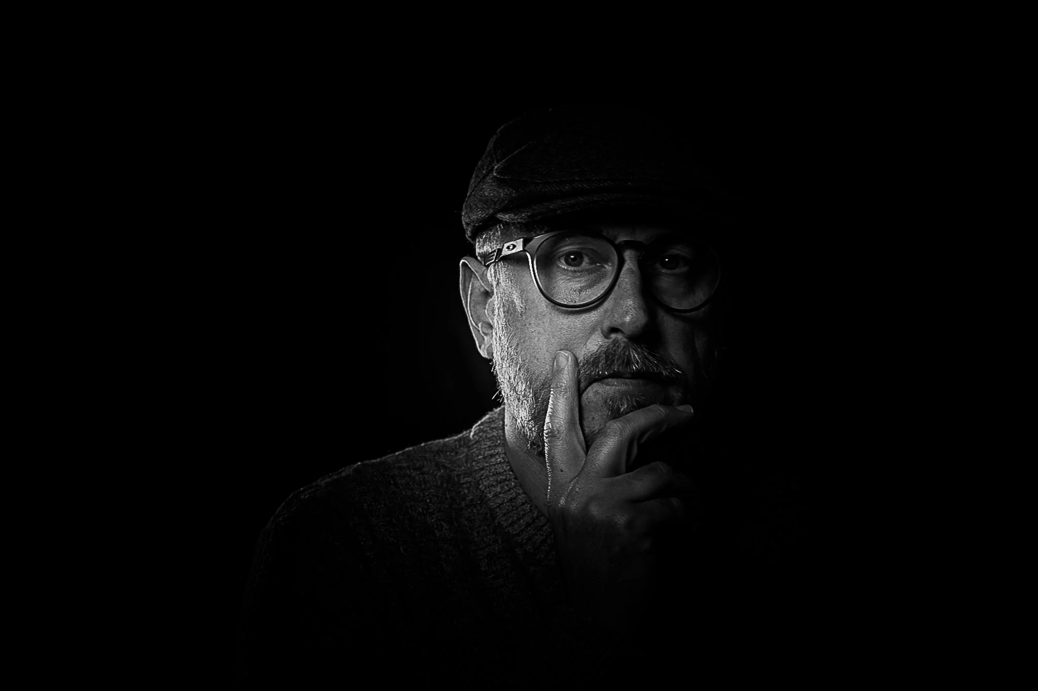 Black and white photo of a man with glasses and a cap, touching his lips with his hand, against a dark background.