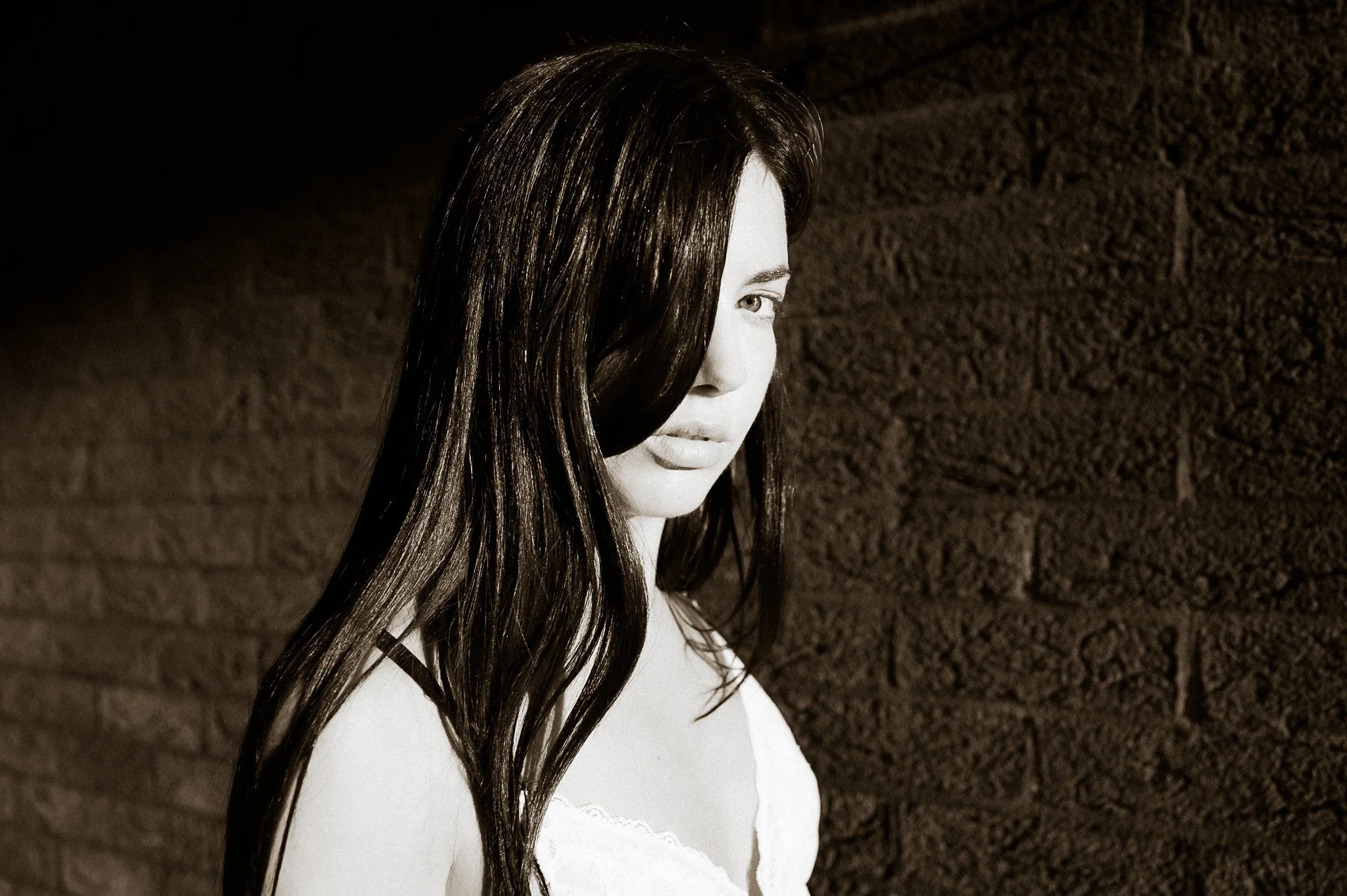 A black and white photo of a woman with long, dark hair, partially covering her face, standing against a brick wall.