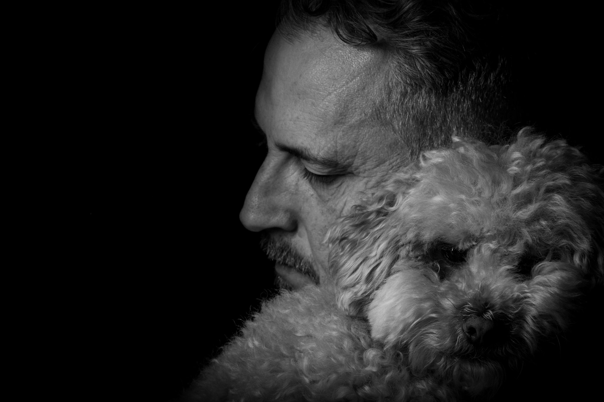 A man with short hair and a beard holding a curly-haired dog close to his face, both appearing calm, in black and white.