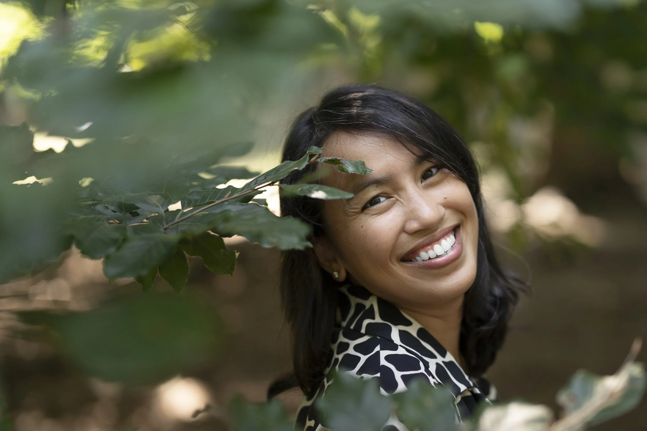 Joyce, smiling and peeking through green leaves in a natural outdoor setting. Photo credits to Nick Fried for Here, Aware website.