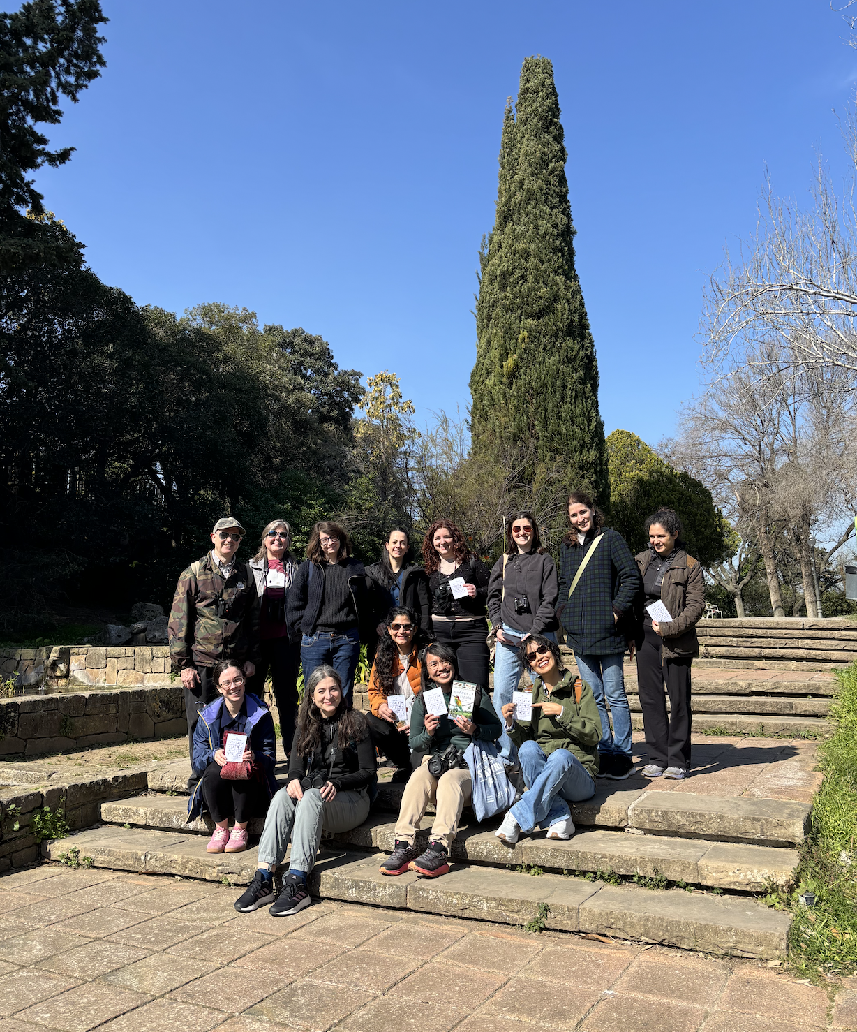 Group of people smiling outdoors on stone steps with a tall cypress tree and greenery in the background.