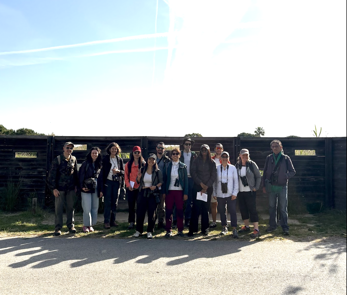 Group of 14 people standing outdoors in front of a black wooden fence on a sunny day, some holding cameras and papers, with clear blue sky overhead.