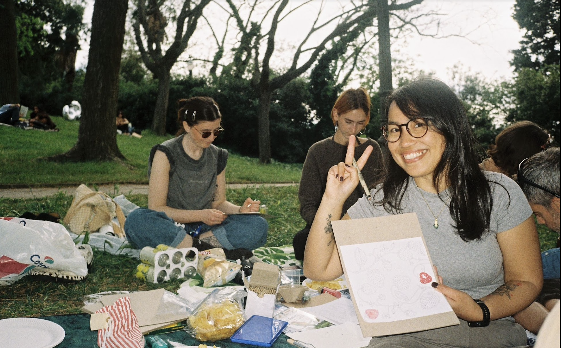 A group of women enjoying a picnic outdoors in a park with trees, sitting on a blanket with food and drinks, one woman smiling and showing a peace sign.