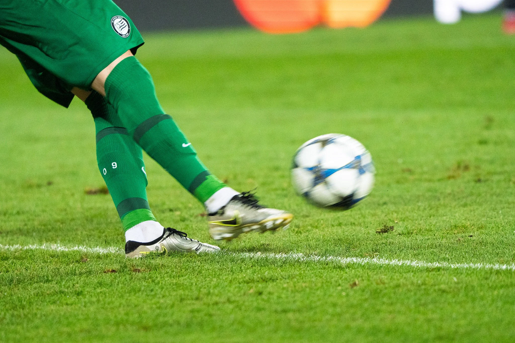 Fußballspieler in grüner Uniform beim Kicken, nur die unteren Beine sichtbar, Fußball im Flug Bewegung auf grünem Rasen. 