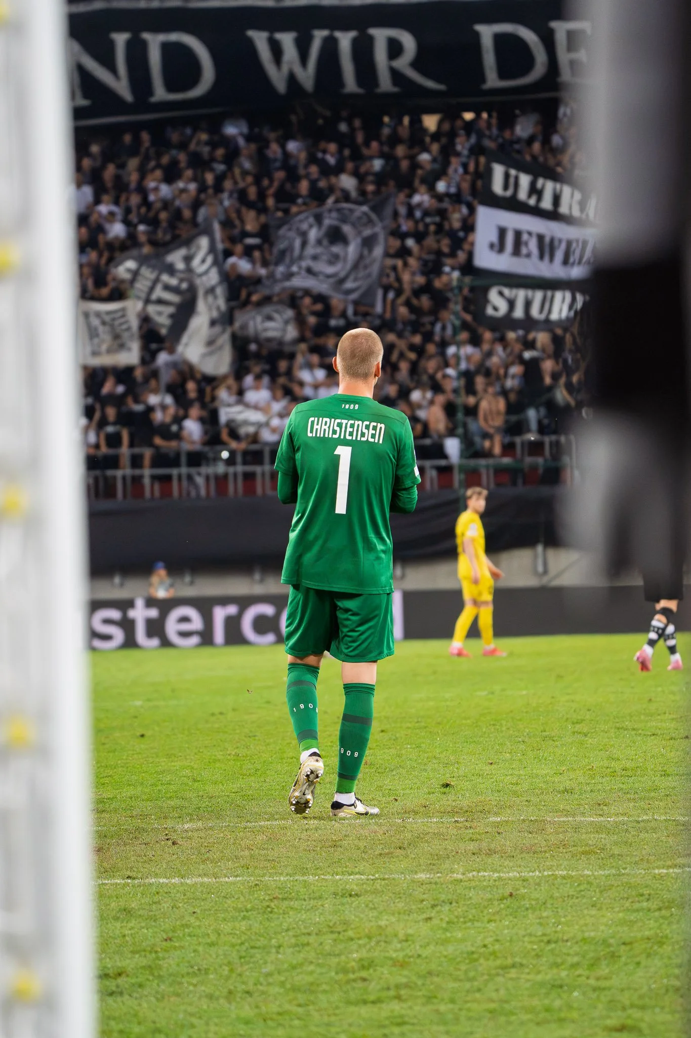 Fußballspieler in grüner Jacke mit der Rückennummer 1 und dem Namen 'Christensen' auf dem Spielfeld während eines Spiels, mit einem Publikum im Hintergrund.