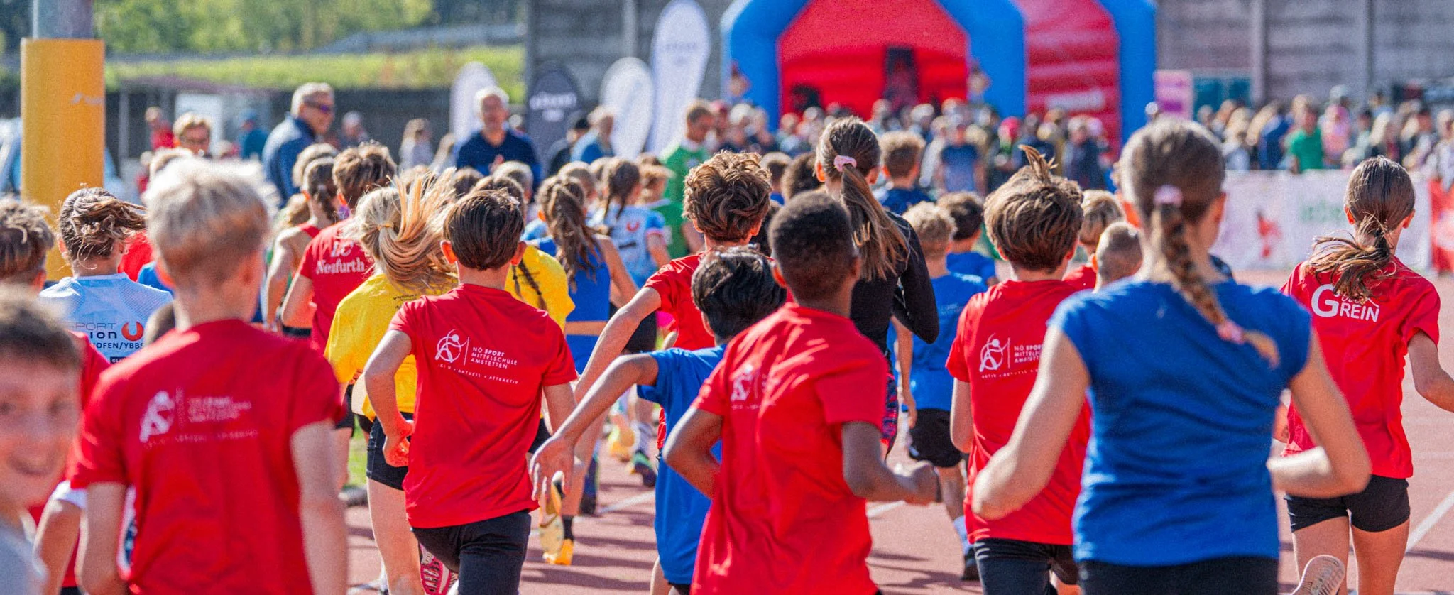 Viele Kinder laufen beim Start eines Laufwettbewerbs auf einer Laufbahn im Freien, alle tragen bunte T-Shirts mit Aufdrucken.