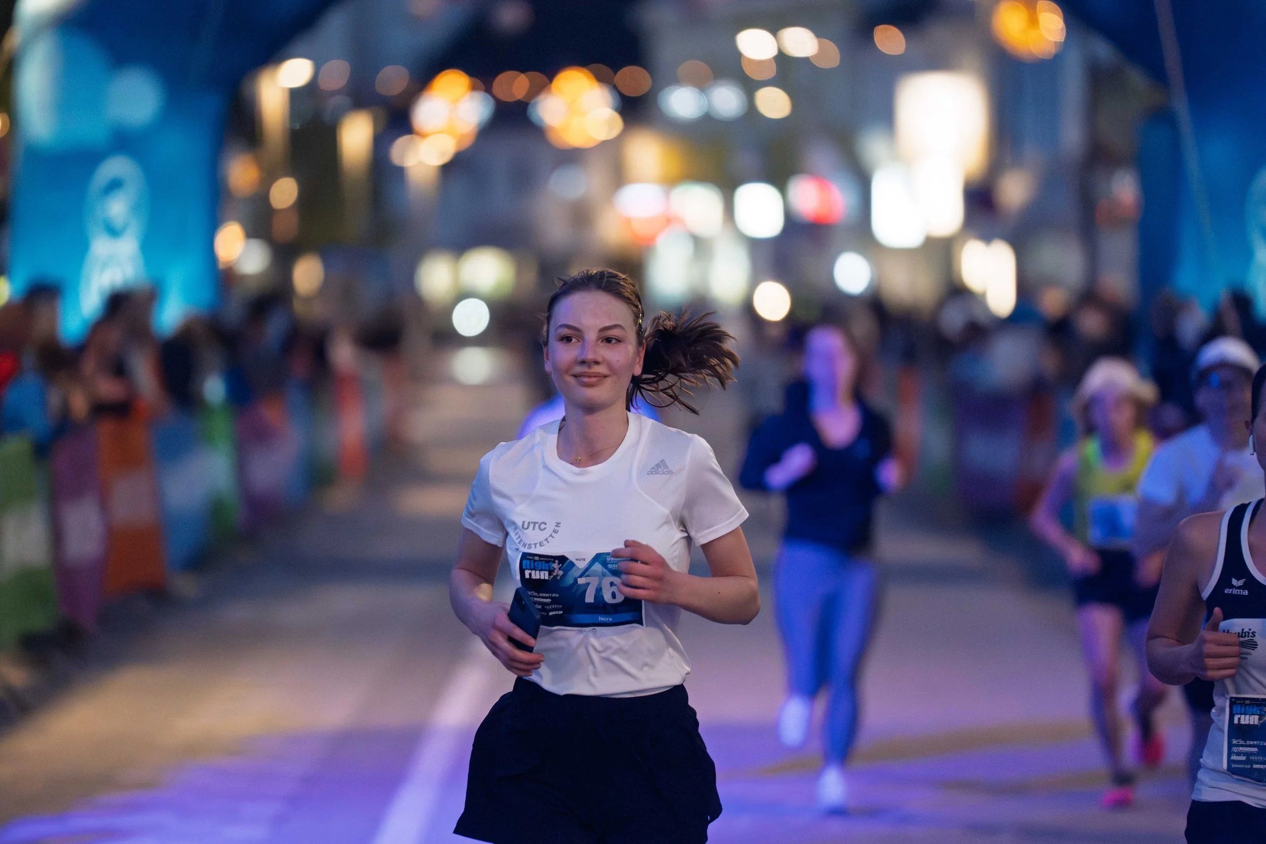 Junge Läuferin bei einem nächtlichen Lauf, umgeben von anderen Teilnehmern und bunten Lichtern im Hintergrund.