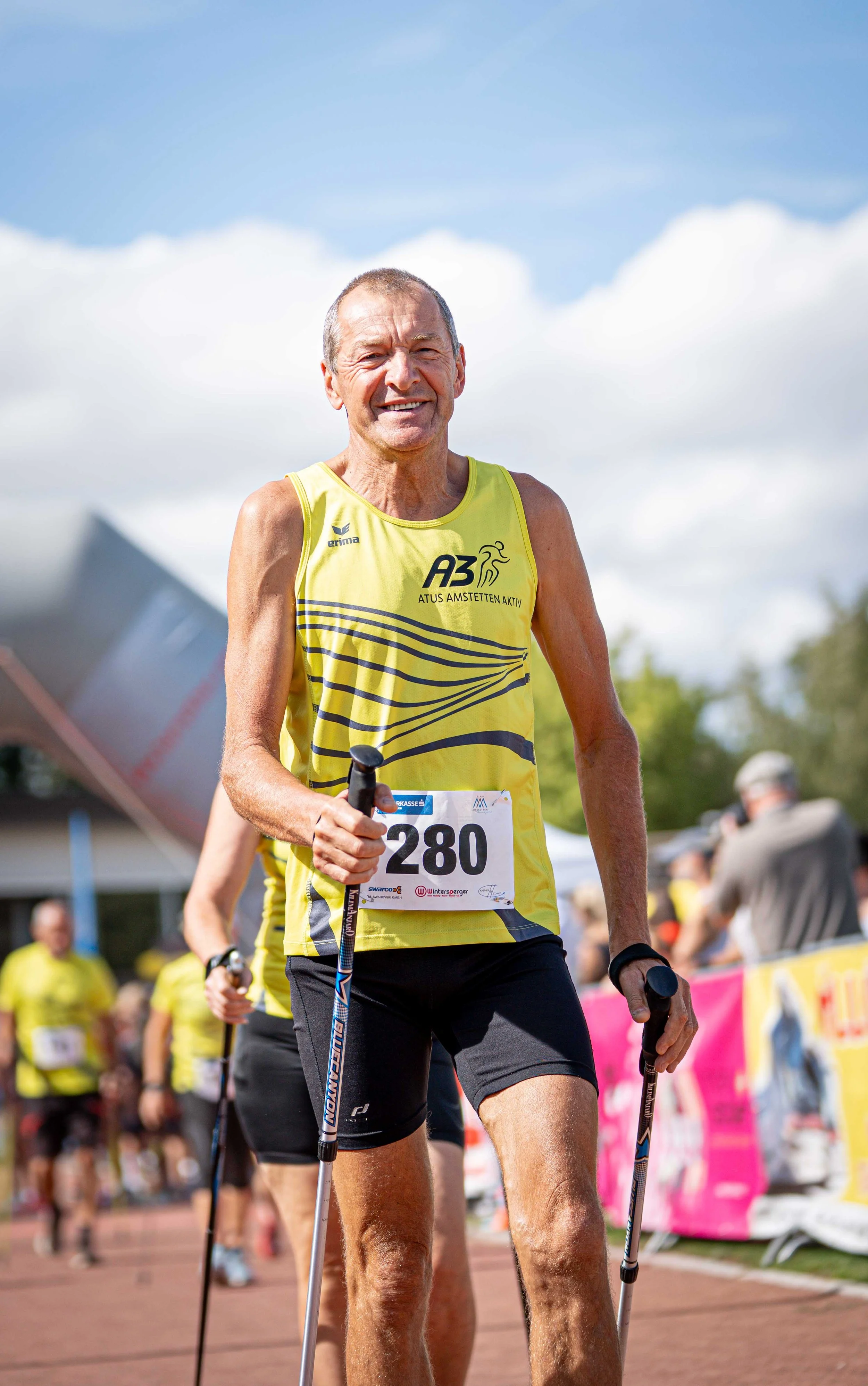 Ein älterer Mann beim Laufwettbewerb, trägt eine gelbe Laufjacke mit der Nummer 280 und benutzt Nordic-Walking-Stöcke. Im Hintergrund sind mehrere Menschen, wahrscheinlich Teilnehmer und Zuschauer, und es ist ein sonniger Tag.