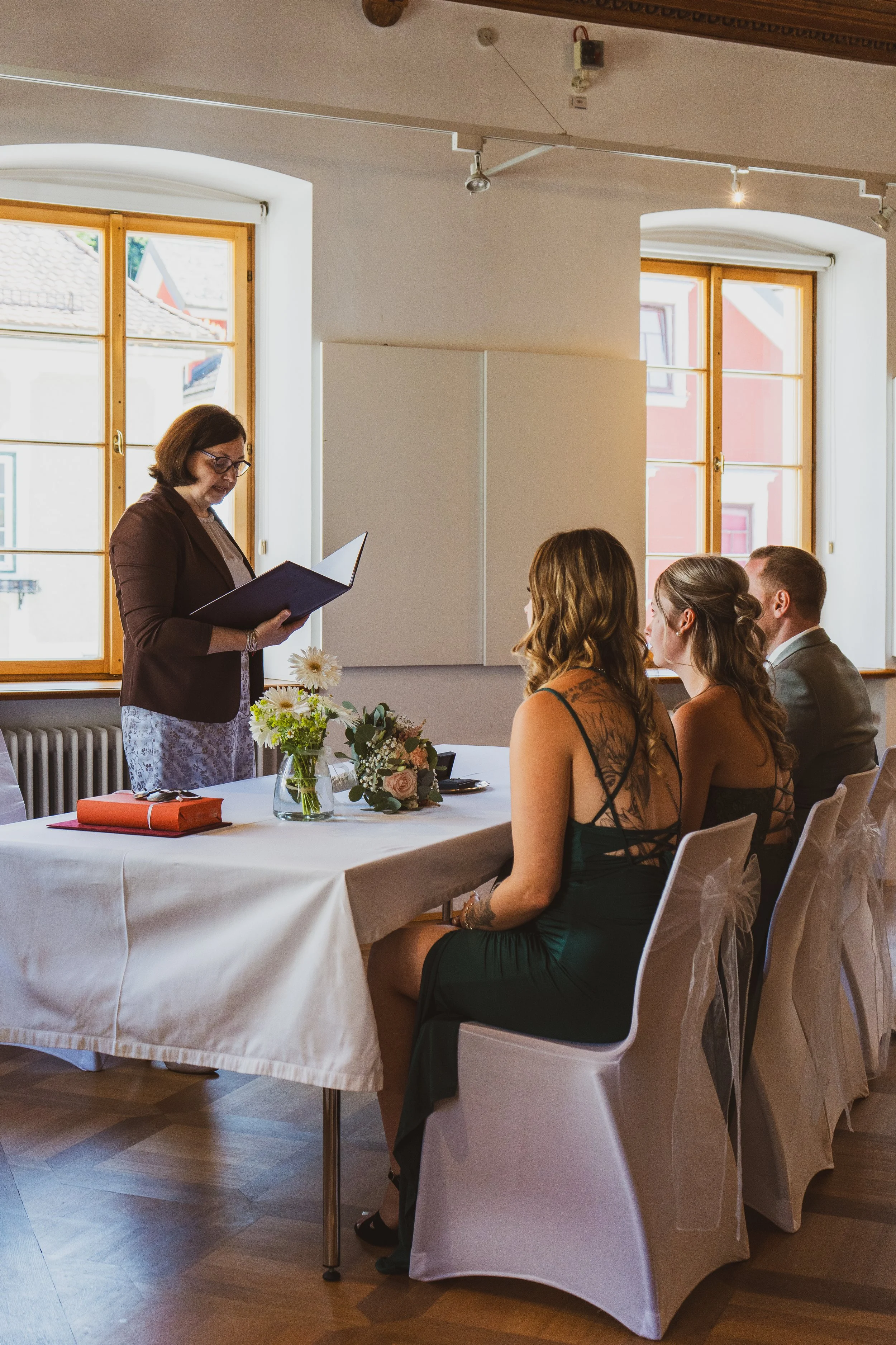 Eine Frau liest während einer Zeremonie für vier Personen einen Text vor. Es sitzen zwei Frauen und ein Mann an einem festlich gedeckten Tisch, der mit Blumen dekoriert ist, in einem Raum mit großen Fenstern.