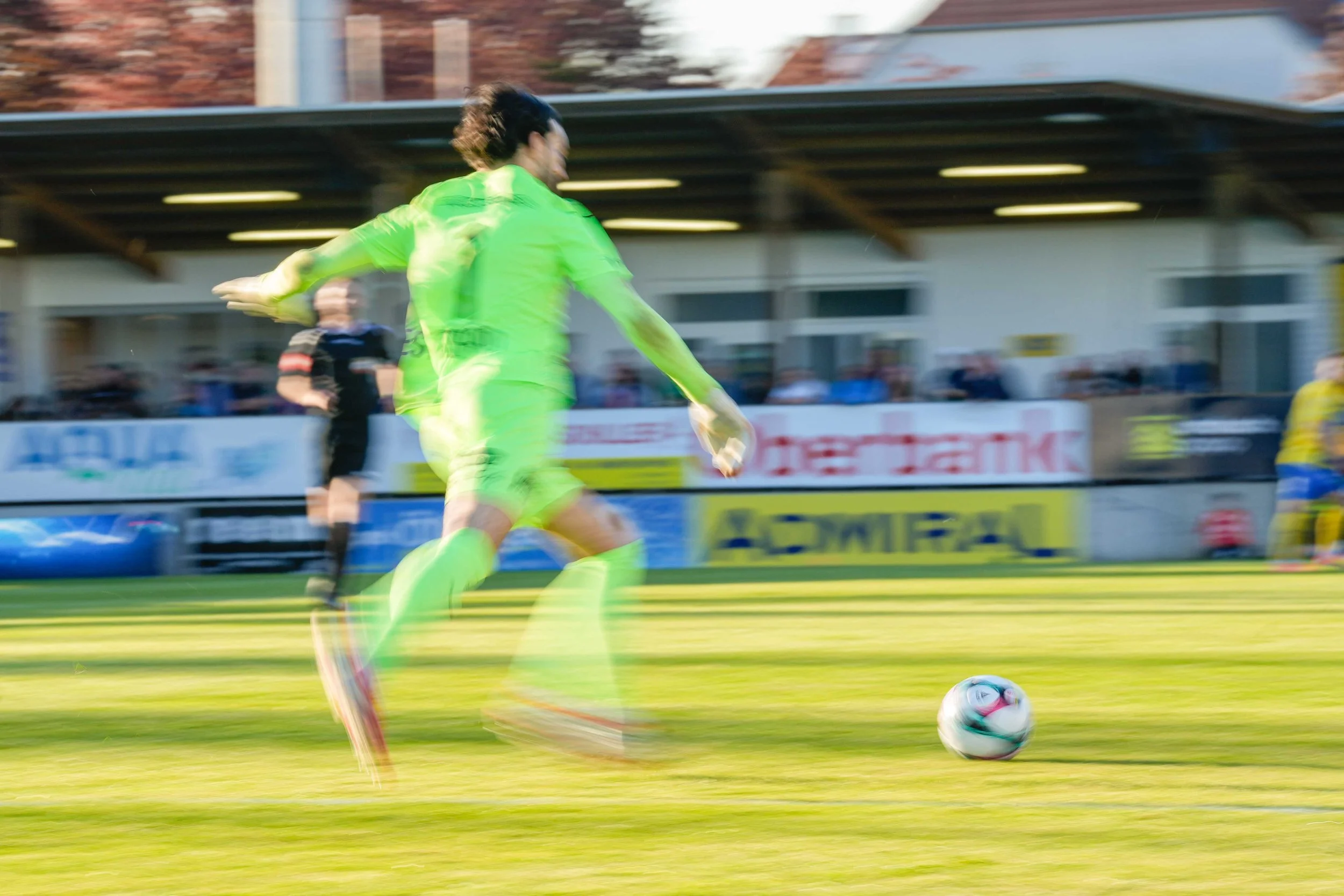 Ein Fußballspieler in Neon-Green beim Schießen eines Balls auf dem Fußballfeld, im Hintergrund ein Publikum im Stadion, bei Dämmerlicht, schnelle Bewegung erkennbar.