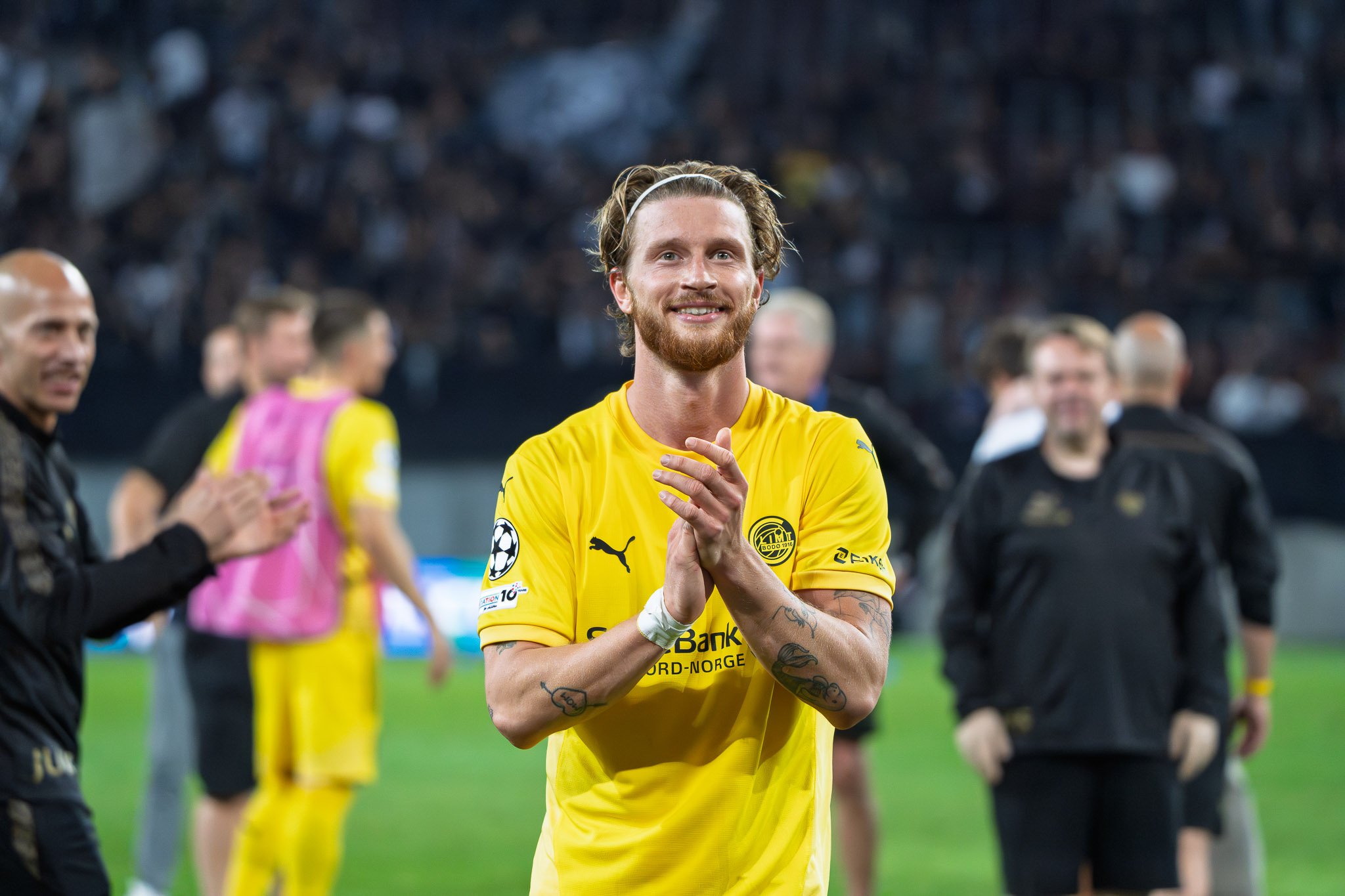 Fußballspieler in gelbem Trikot nach einem Spiel, klatscht und lächelt, im Stadion, mit anderen Teammitgliedern im Hintergrund.