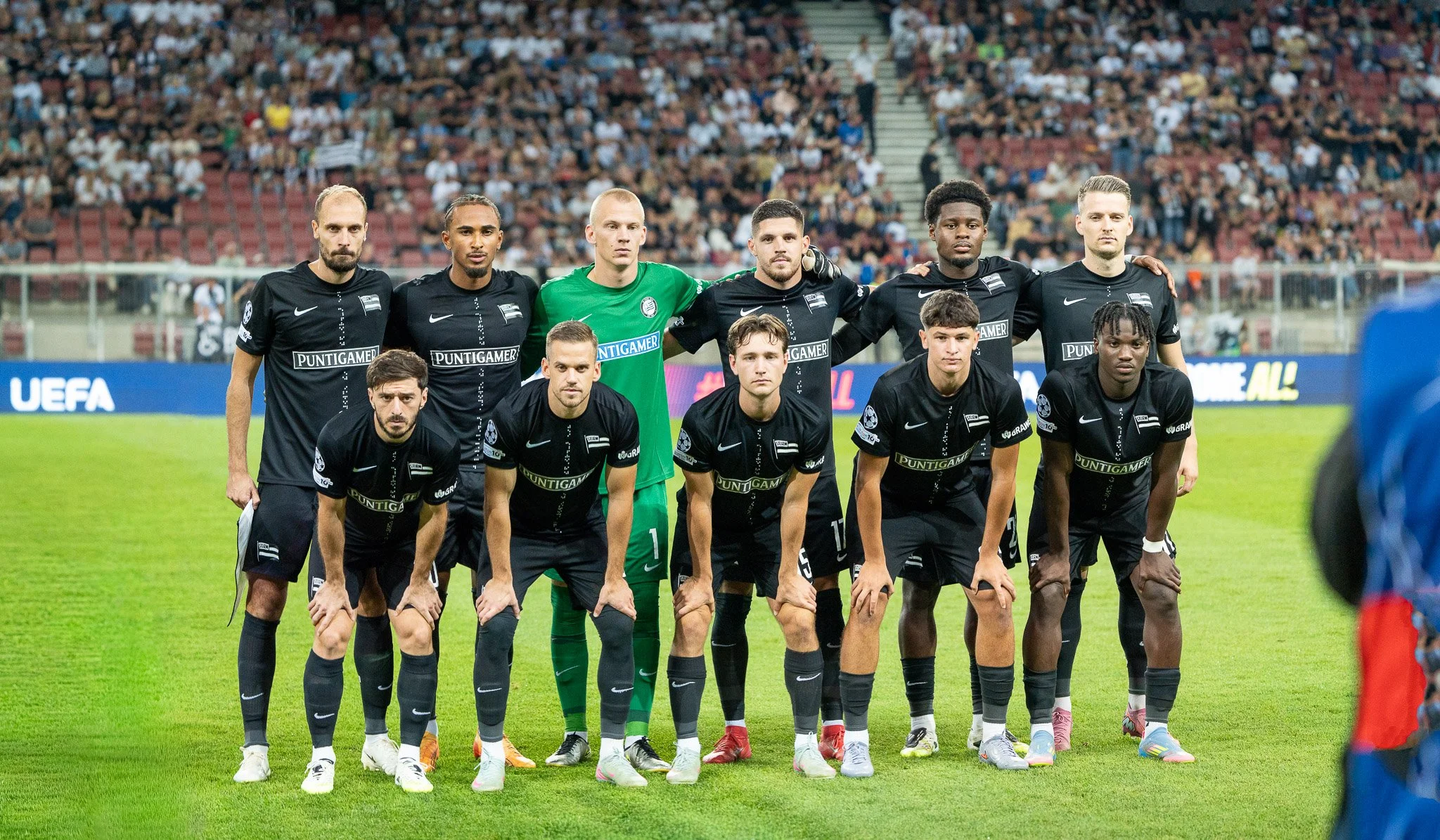 Fußballteam posiert für ein Mannschaftsfoto auf dem Spielfeld vor Zuschauern im Stadion.