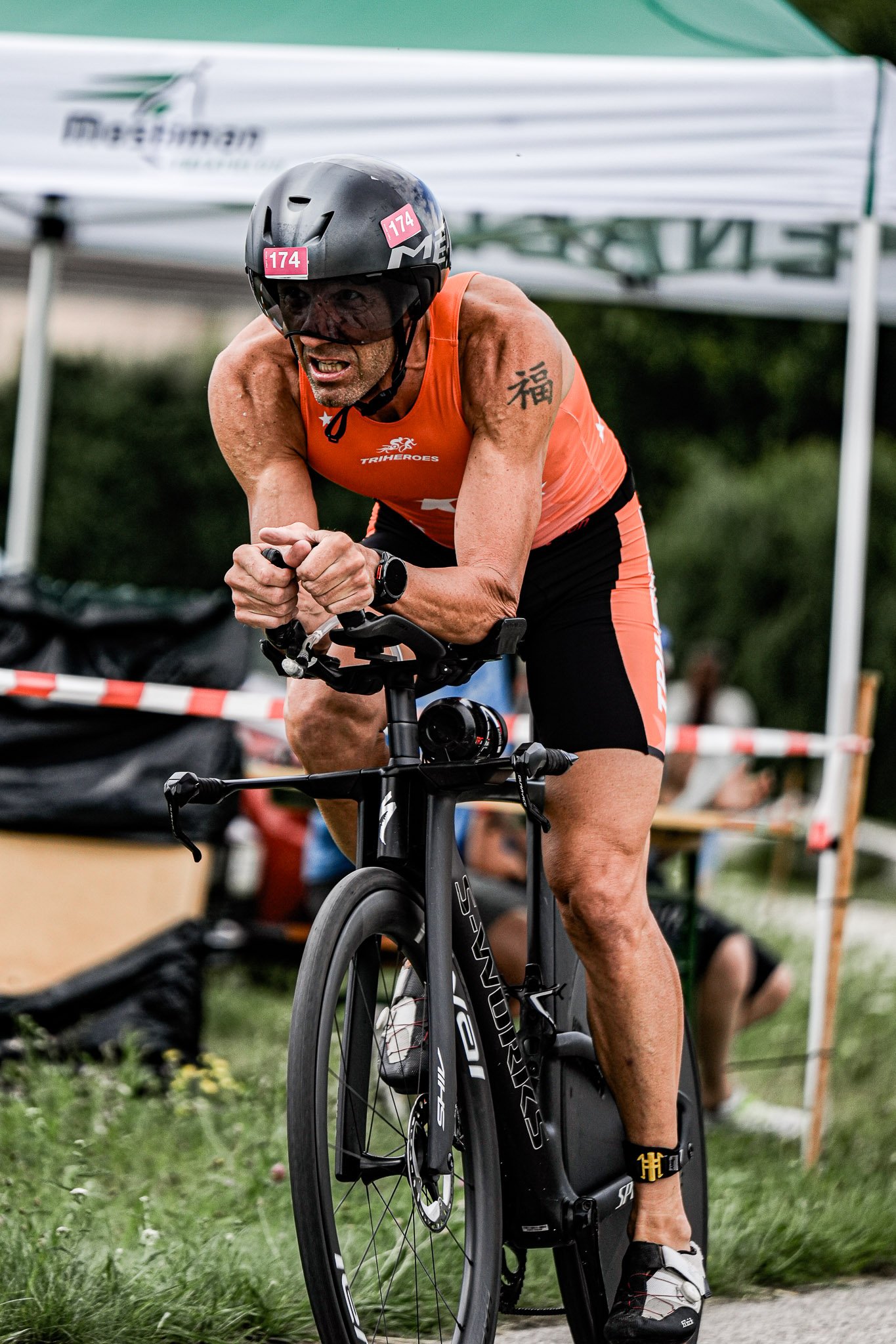 Ein Mann beim Triathlon auf einem schwarzen Rennrad, trägt einen schwarzen Helm, eine orange Weste und schwarze Fahrradhosen, während er an einem Wettkampf teilnimmt.