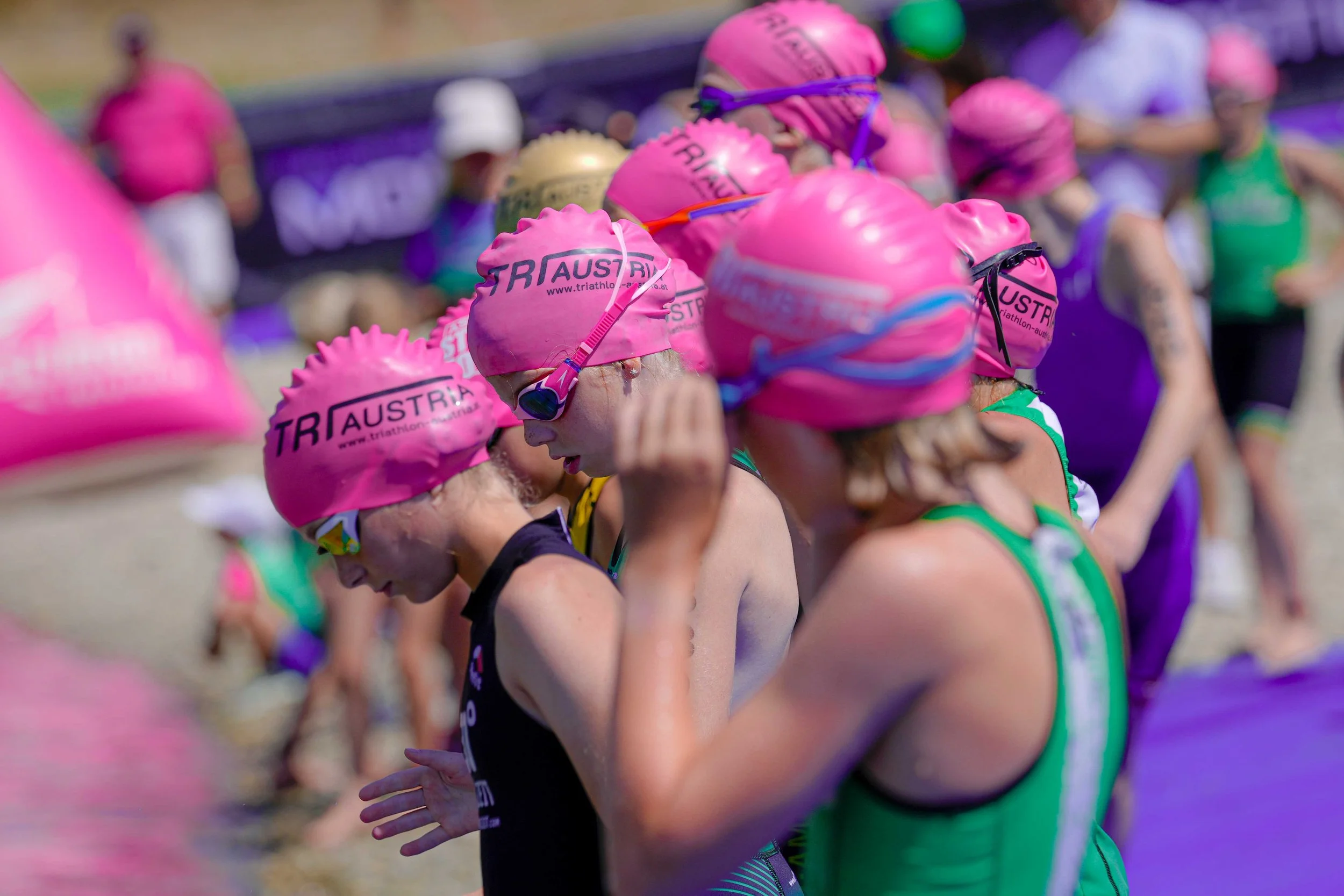 Mehrere Kinder beim Triathlon, alle tragen pinke Badekappen mit der Aufschrift 'TRI AUS TRIA' und Schwimmbrillen. Sie scheinen sich auf den Start oder das Rennen vorzubereiten.