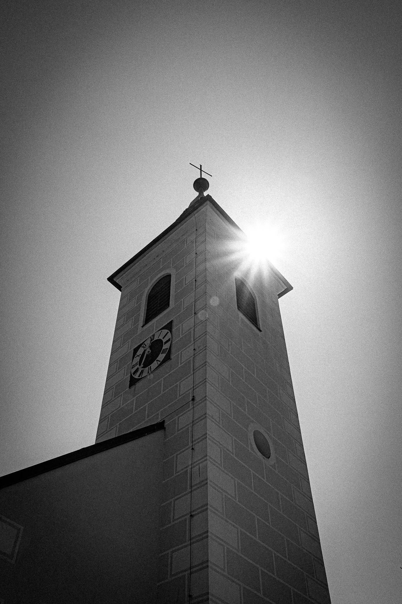 Schwarz-weiß-Foto eines Kirchturms mit Uhr, Sonnenstrahlen hinter dem Turm, Himmel im Hintergrund.