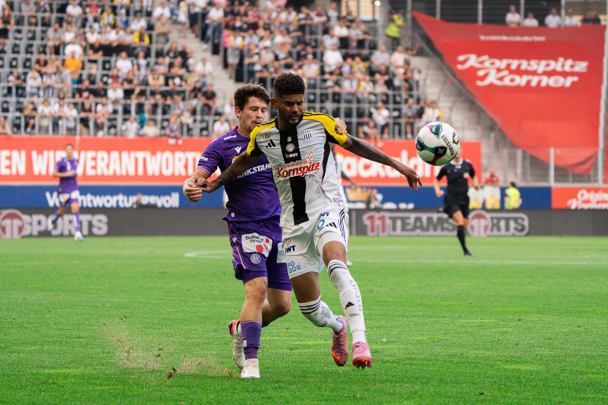 Zwei Fußballspieler kämpfen um den Ball auf dem Spielfeld, im Hintergrund sind Zuschauer im Stadion. Ein Schiedsrichter ist sichtbar.