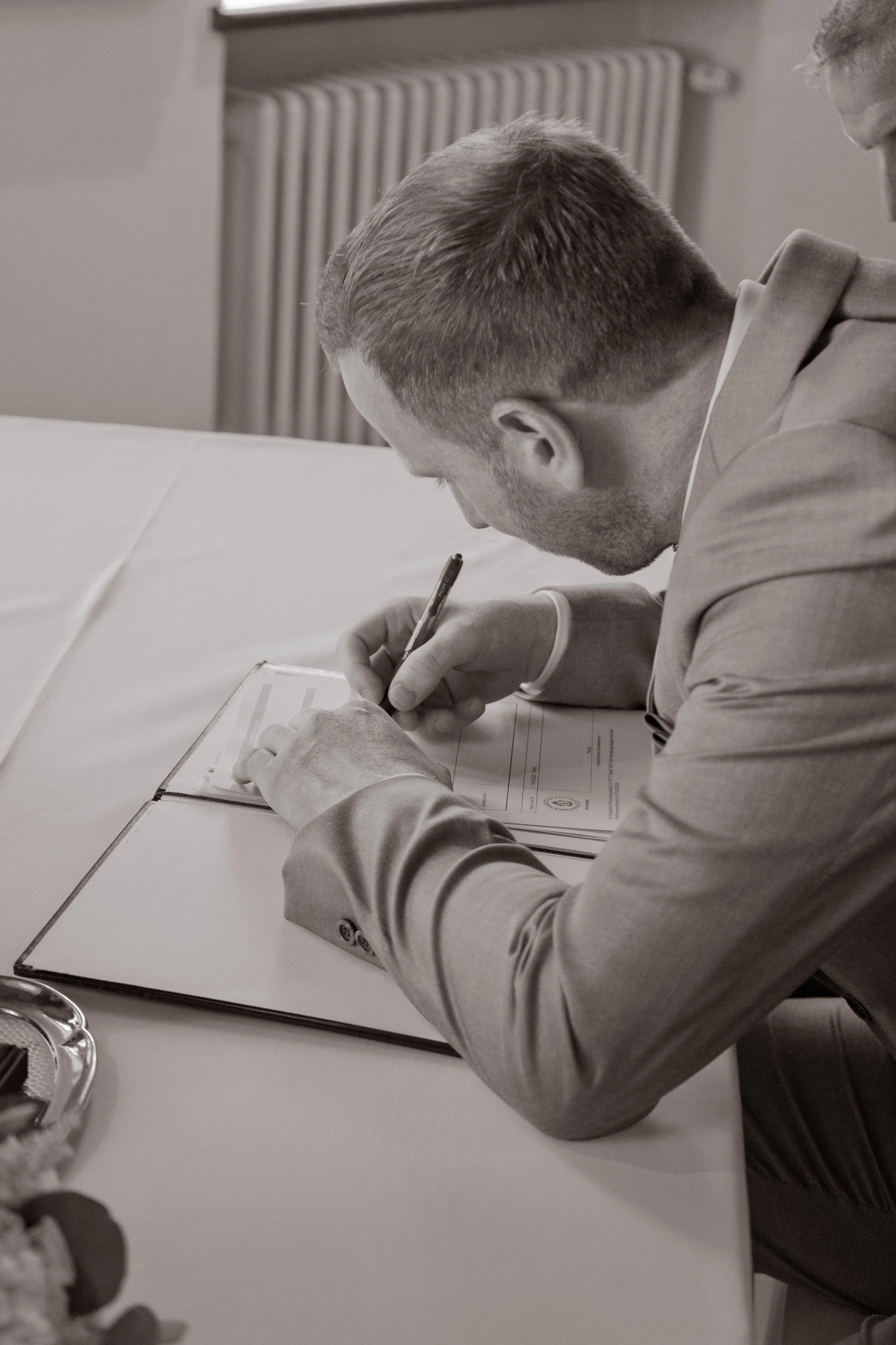 Ein Mann in einem Anzug unterschreibt ein Dokument auf einem Tisch in einem Raum mit Heizkörper im Hintergrund.