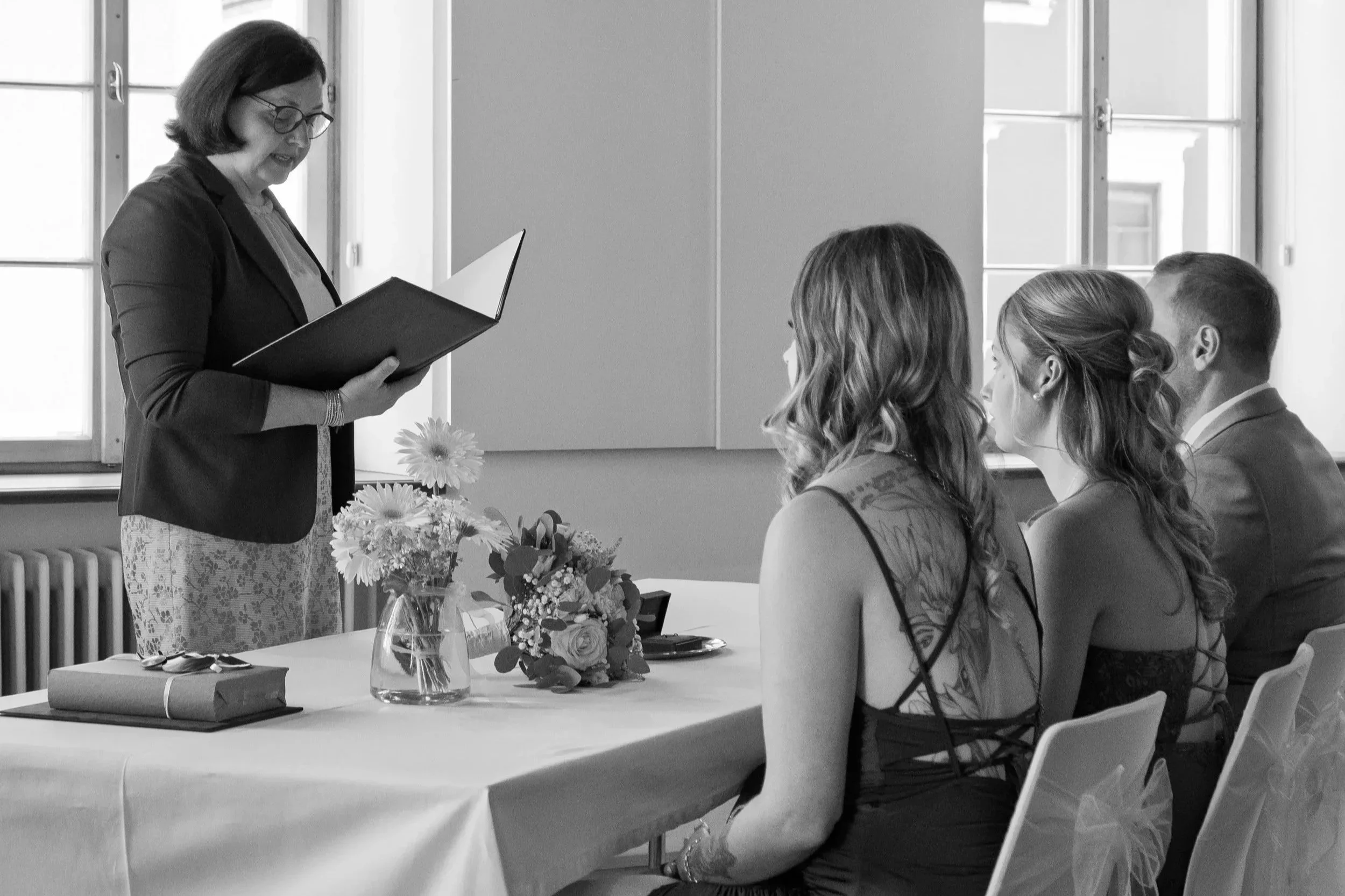 Eine Frau liest vor einer Gruppe von drei Menschen in einem Raum, Tische und Blumen sind sichtbar.