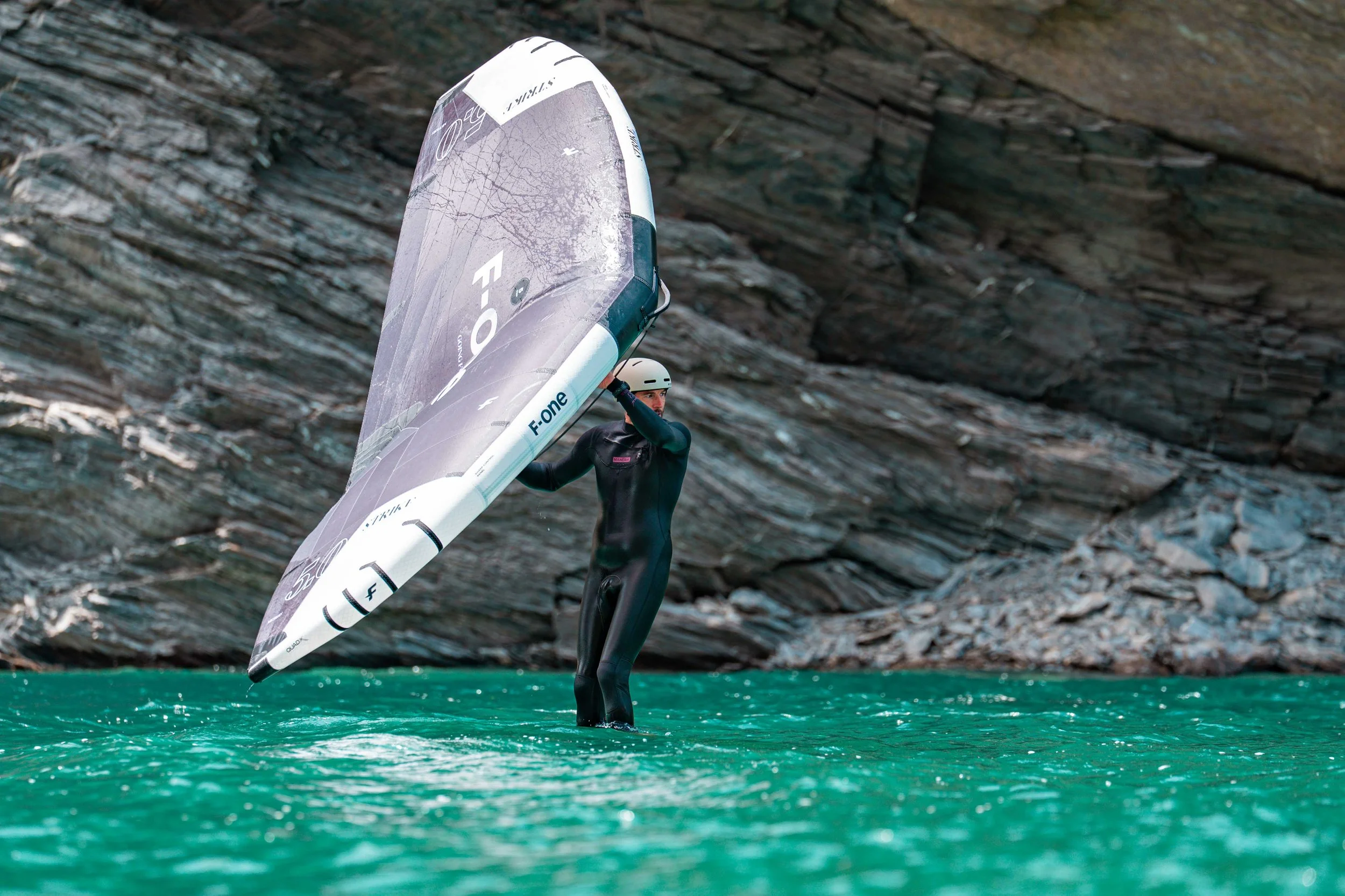 Ein Segler in einem Neoprenanzug trägt ein Windsurfing-Board in der Hand und steht im Wasser, vor einer steilen, felsigen Klippe.