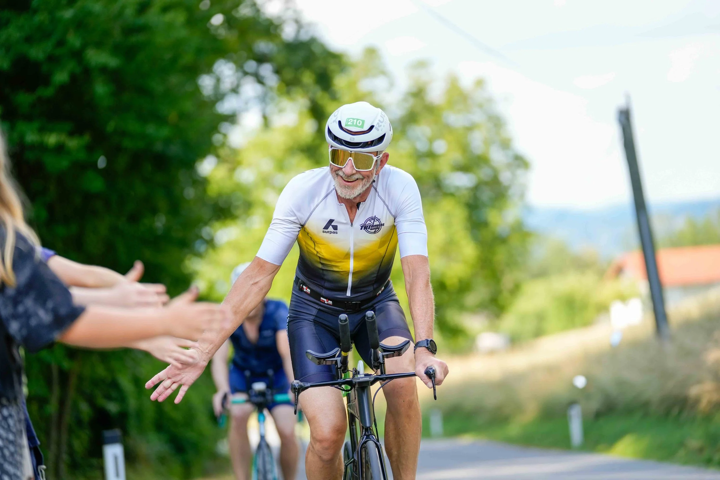Ein männlicher Radfahrer, der lächelnd einen Handshake mit einer Person am Straßenrand macht. Der Radfahrer trägt einen Helm und Sonnenbrille. Es ist eine grüne, sonnige Umgebung.