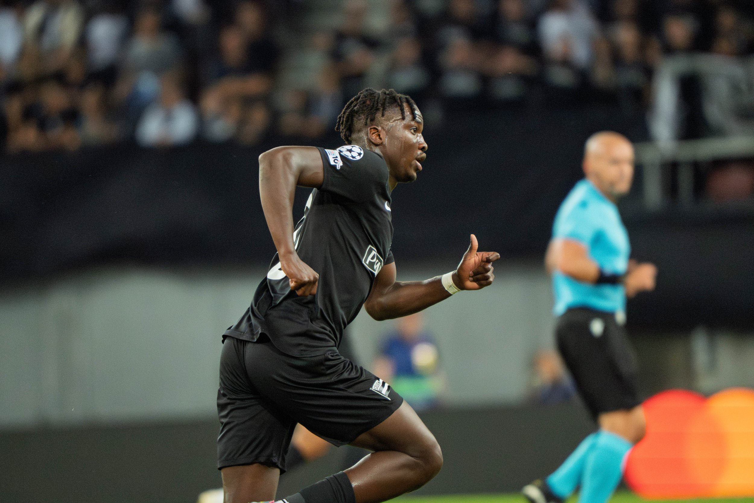 Fußballspieler in Schwarz während eines Spiels auf dem Spielfeld, mit einem Schiedsrichter im blauen Shirt im Hintergrund.