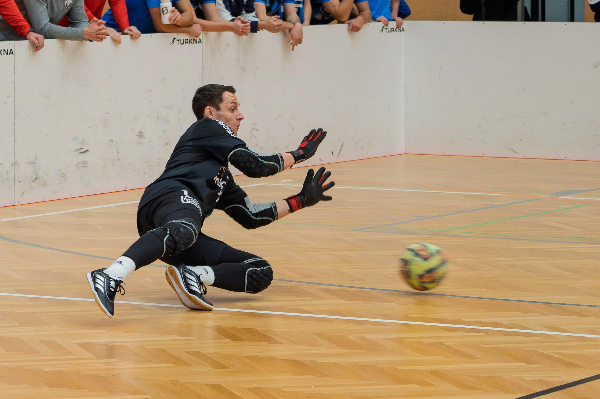 Fänger im Blaumann im unteren Bild, beim Fußball im Indoor-Sportfeld, der gerade den Ball abfängt.