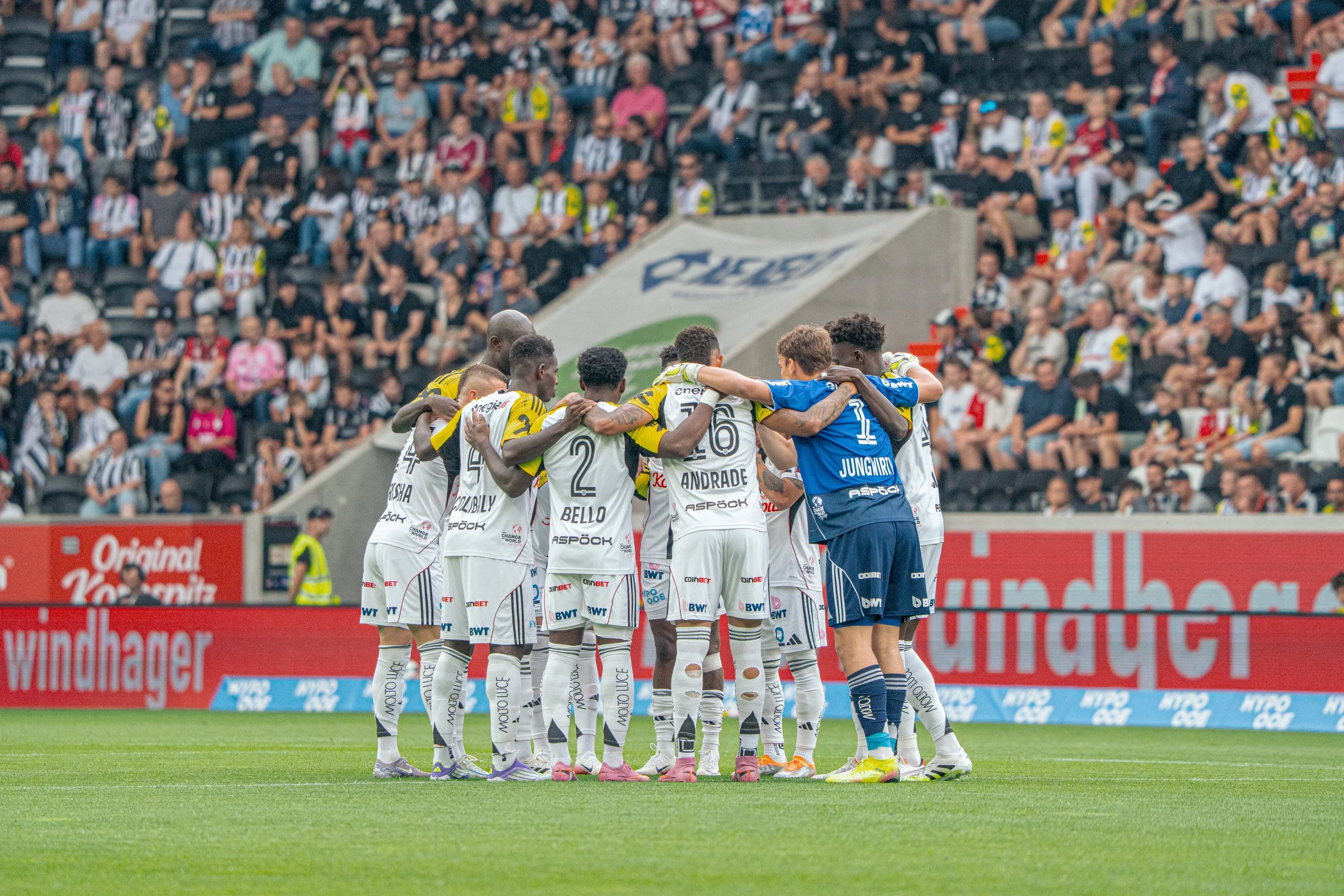 Fußballspieler in weißen Trikots und blauen Torwarttrikots versammeln sich in einer Team-Huddle auf dem Spielfeld, während im Hintergrund eine große Zuschauermenge im Stadion sitzt.