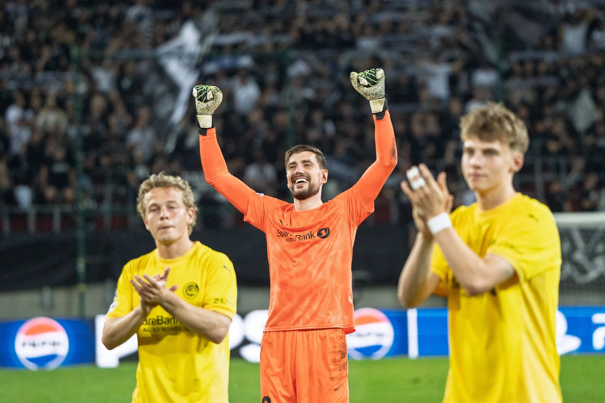Fußballtorwart in orangeem Trikot mit ausgebreiteten Armen und lachendem Gesicht, umgeben von zwei Spielern in gelben Trikots, auf einem Fußballplatz vor einer Zuschauertribüne.
