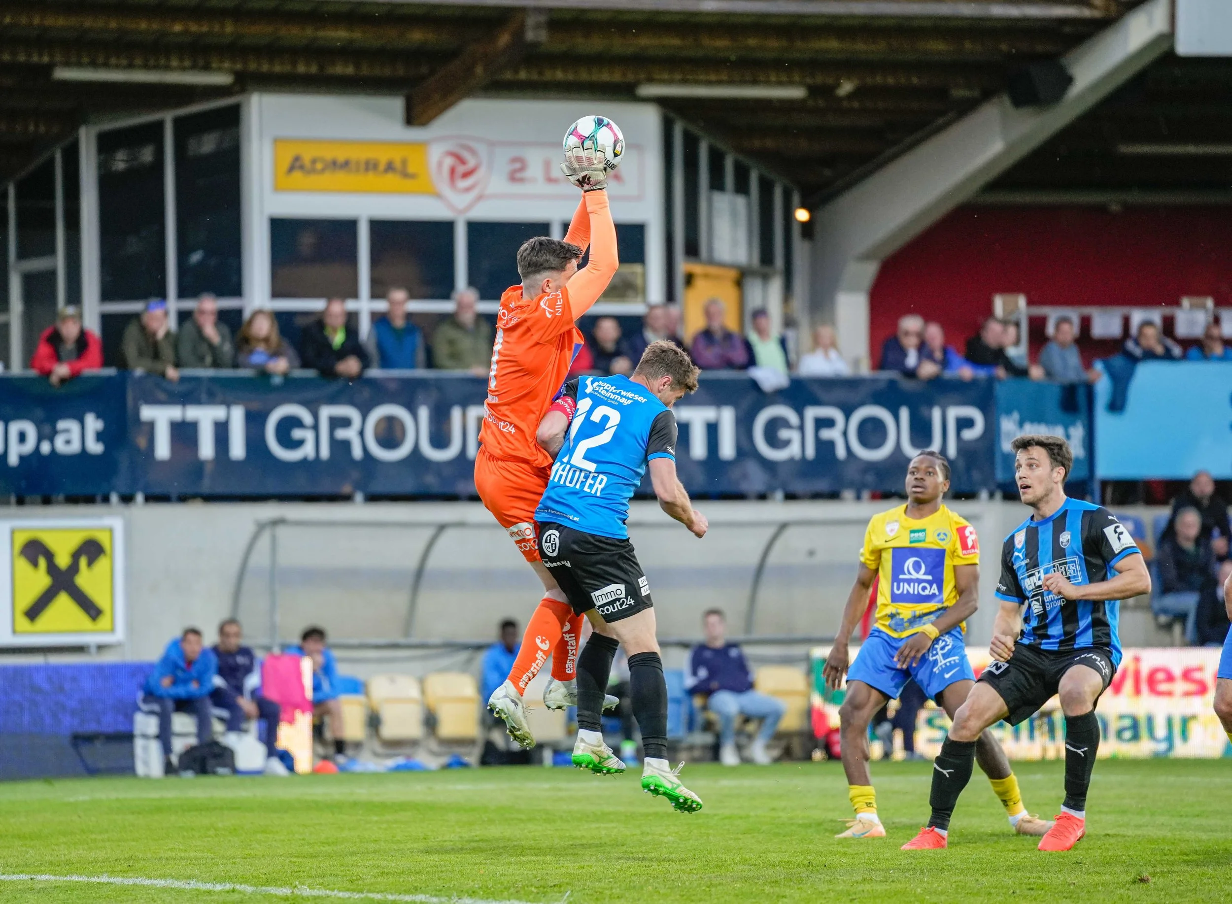 Fußballspiel mit einem Torwart in orange, der hochspringt, um den Ball zu fangen, während mehrere Spieler im blauen und gelben Trikots auf dem Spielfeld sind. Zuschauer sitzen im Hintergrund.