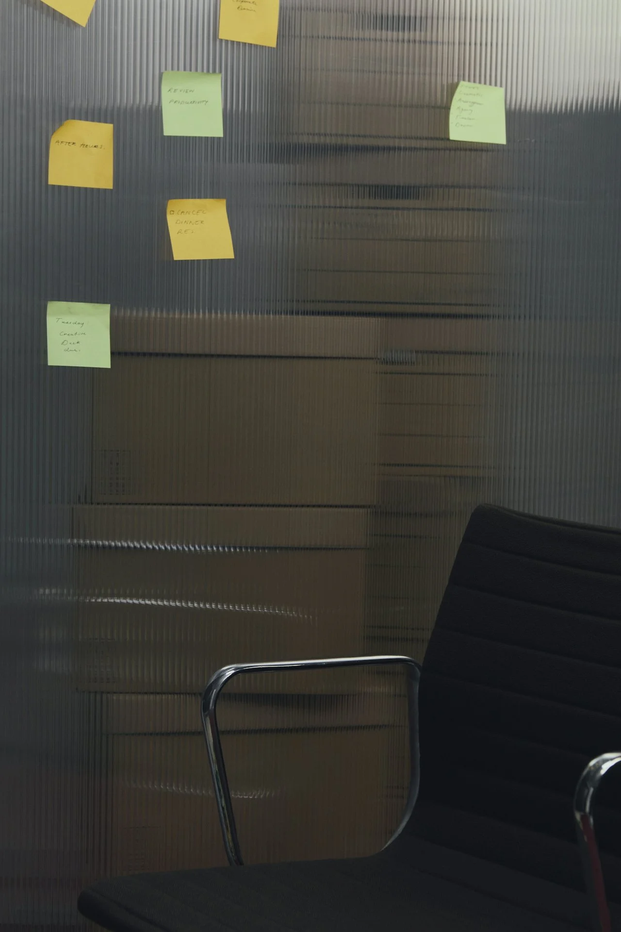 Sicht auf eine Glastür mit mehreren bunten Haftnotizen, darunter grüne und gelbe, mit unterschiedlichen Notizen. Im Vordergrund eine schwarze Büromöglichkeit mit Armlehnen.