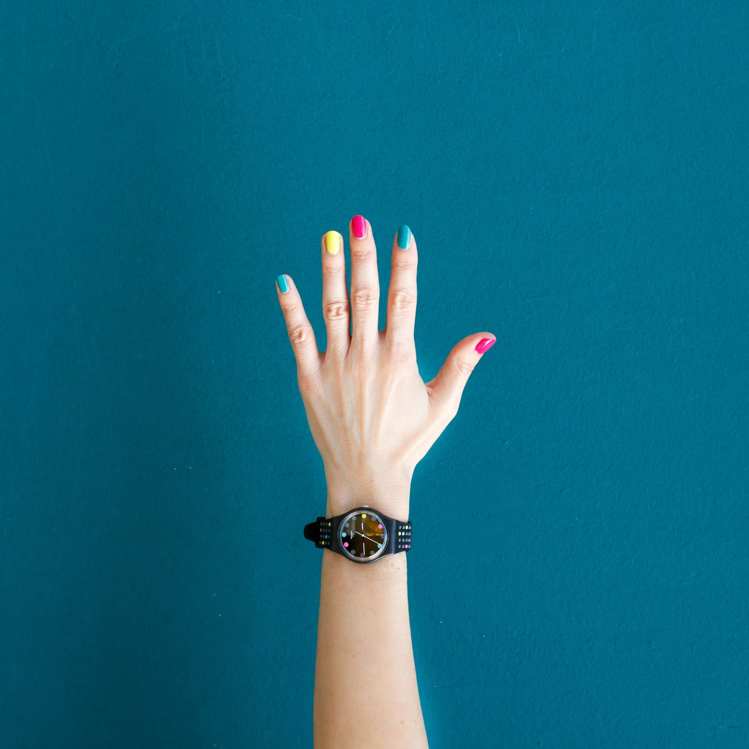 A person's hand is raised against a teal background, showing painted fingernails in yellow, pink, and light blue. The person is wearing a black watch with a colorful dotted face on their wrist.