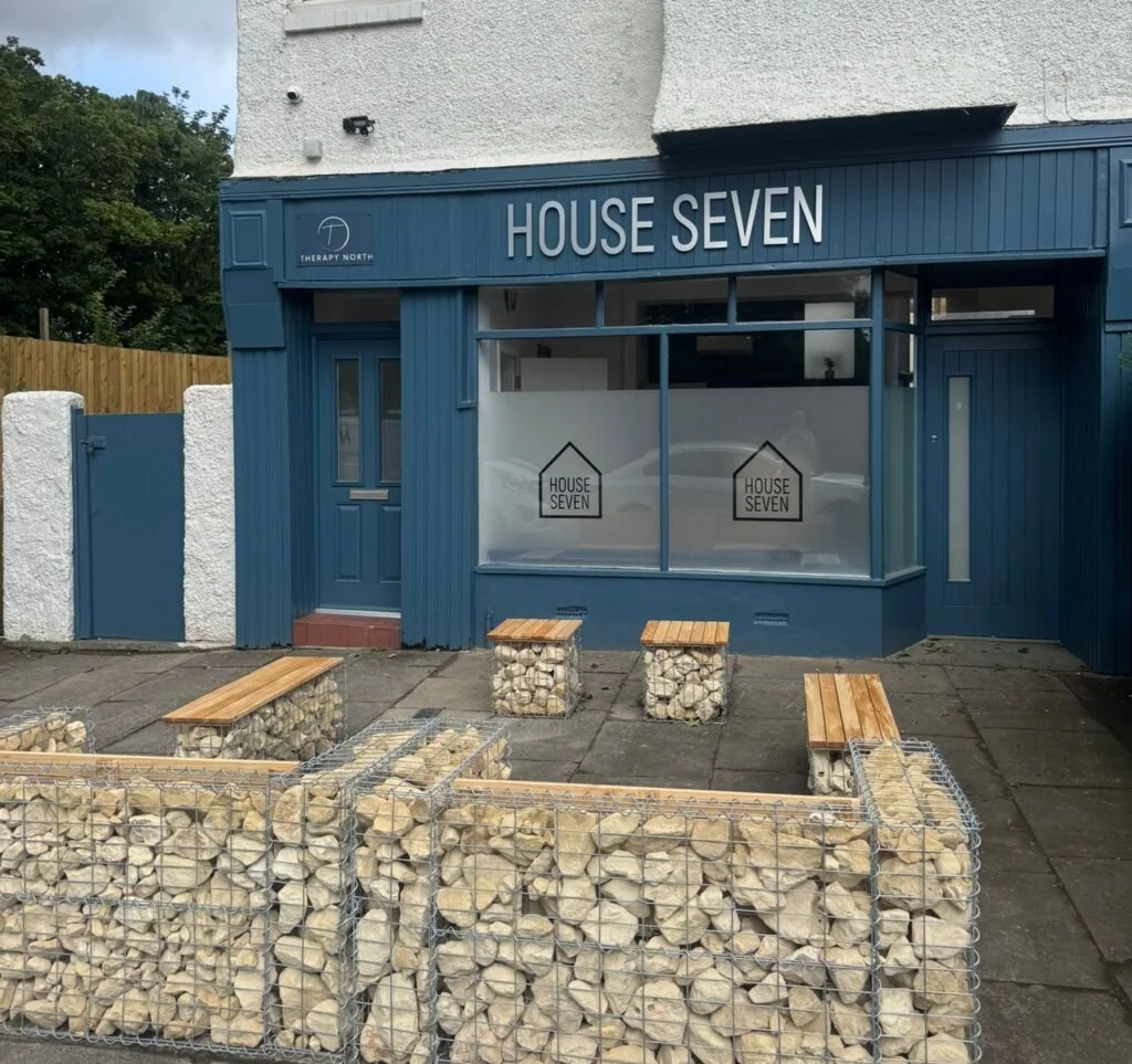 Front of a blue storefront named "House Seven" with large windows and a door, with two benches and stone-filled wire containers in front.