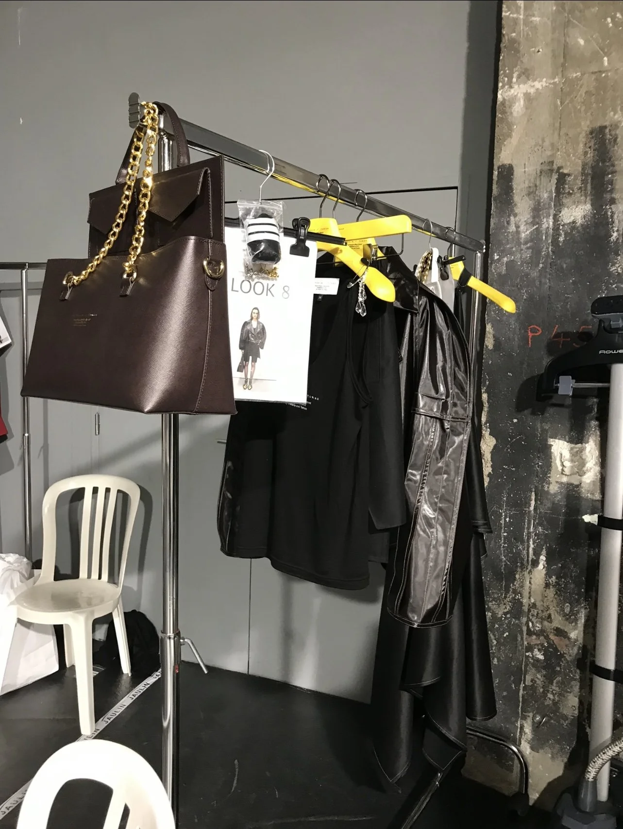 Perchero con bolsas, vestimenta negra, carpetas y accesorios en un entorno de estudio o vestuario, con una pared de fondo de textura industrial.
