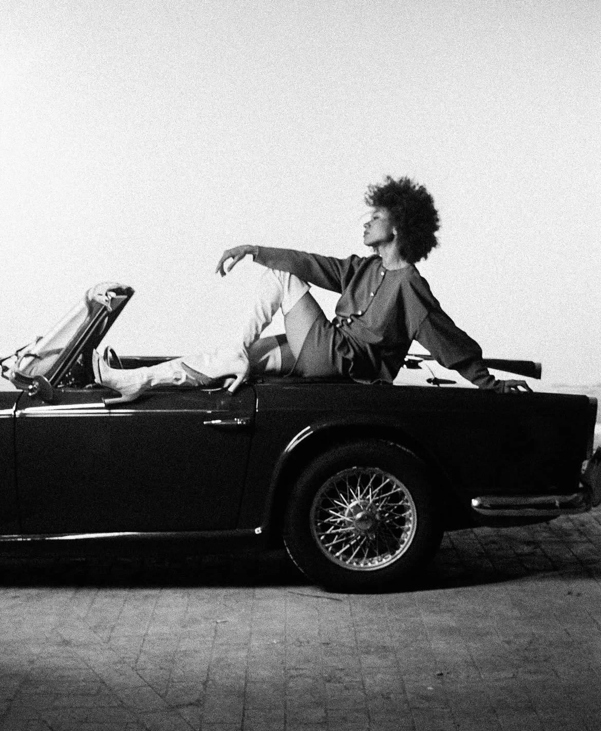 Une femme noire aux cheveux afro repose sur une voiture décapotable en noir et blanc, lunettes de soleil, coiffée d'une robe ou chemise élégante, dans un style artistique et sophistiqué