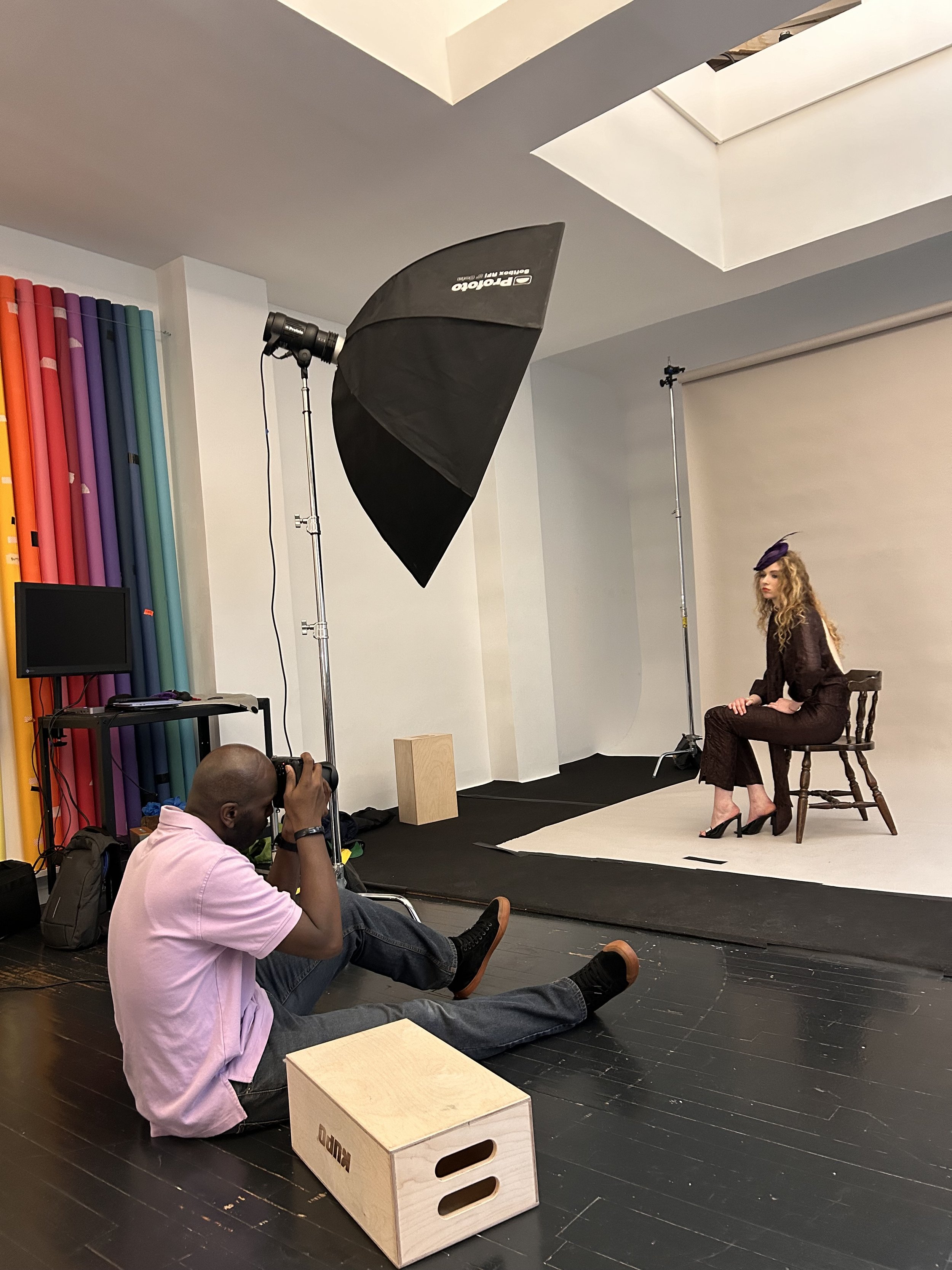 Une séance photo en studio avec un fond blanc, un parapluie de studio, un appareil photo sur trépied et deux personnes, dont une femme élégante assise sur une chaise, et un homme en position assise au sol prenant une photo.