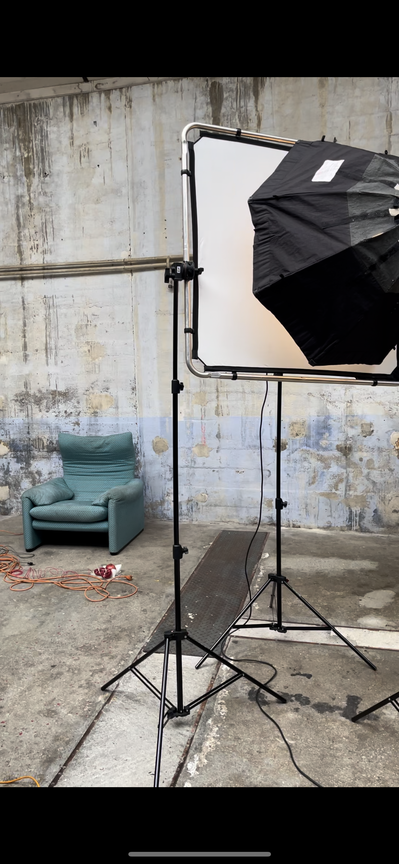Un studio de photographie avec un fond blanc, un parapluie d'éclairage noir et une chaise bleue dans un mur en béton usé.