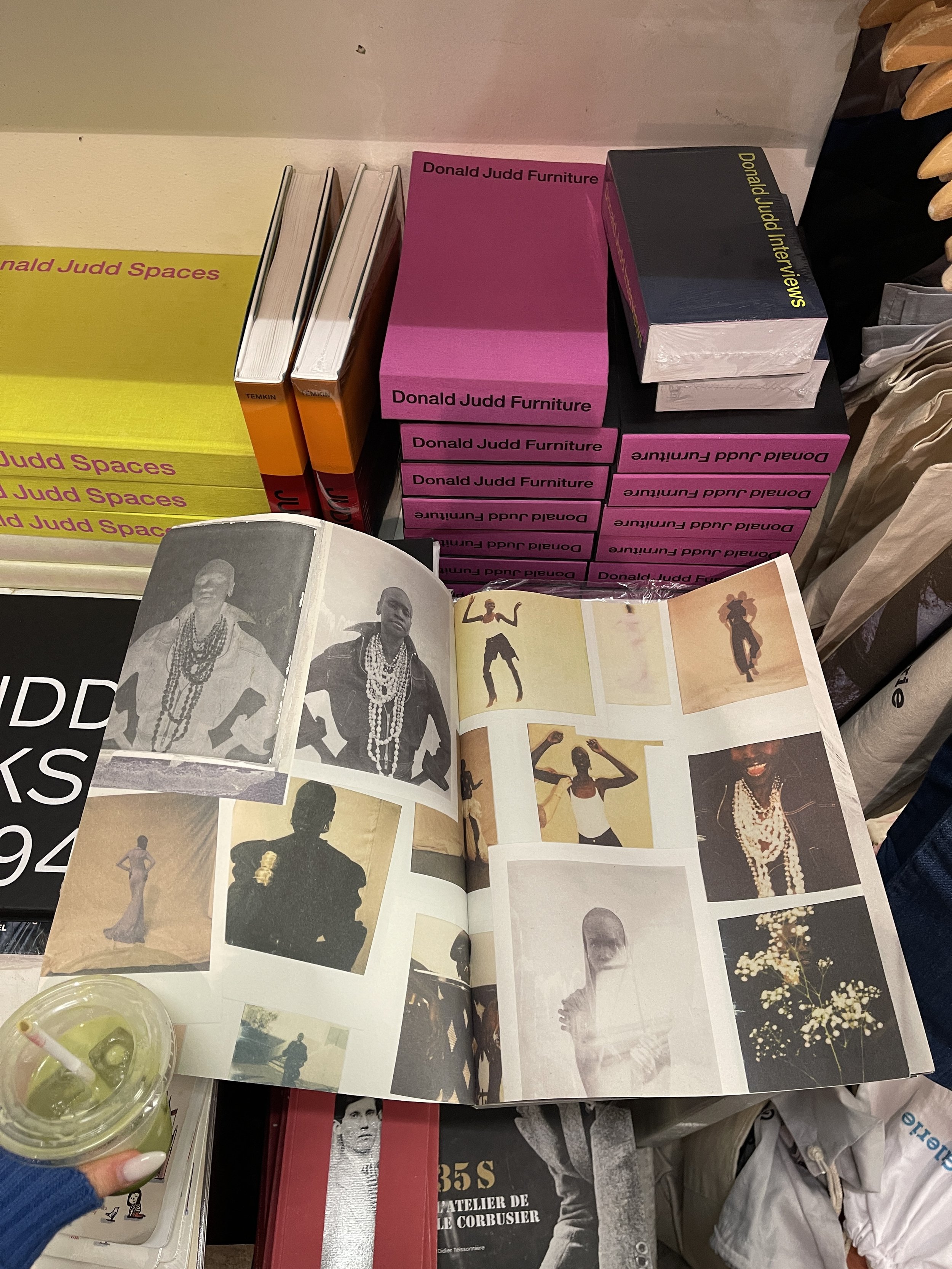 Étals de livres et magazines, avec une personne tenant une boisson verte dans un gobelet en plastique transparent. La table a des magazines ouverts avec des photographies en noir et blanc et en couleur, représentant des personnes et des portraits, ainsi que des illustrations artistiques.