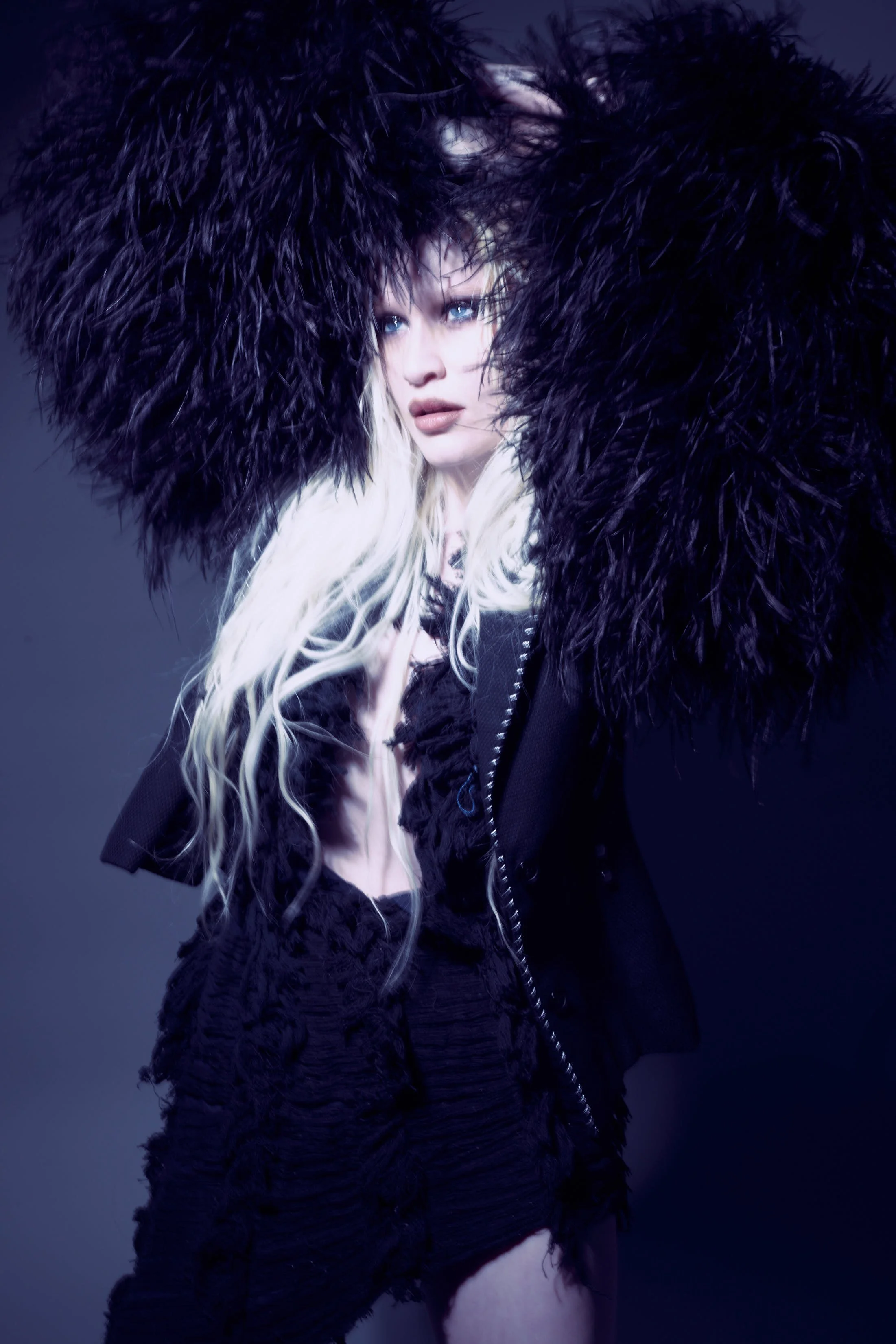 Une femme avec des cheveux longs blonds et des yeux bleus, portant une veste en fourrure noire, regarde vers la gauche, dans un fond sombre.