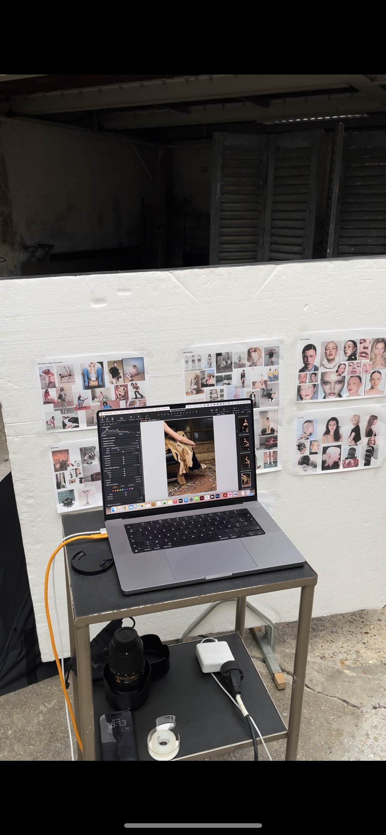 Un ordinateur portable sur une table, entouré de divers appareils électroniques, dans un espace de travail ou un studio avec des photos de modèles affichées en arrière-plan.