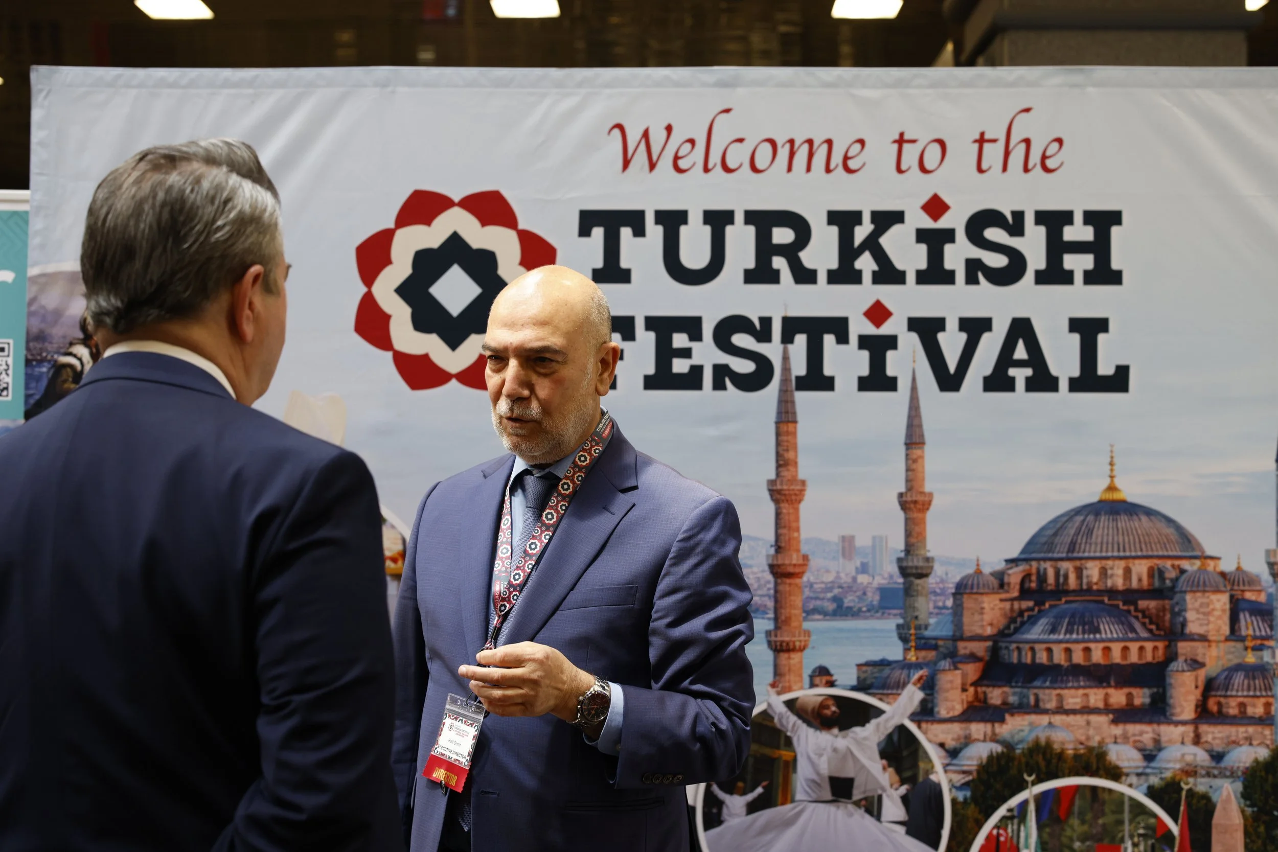 Two men in suits having a conversation at a Turkish Festival, with a banner in the background that reads 'Welcome to the Turkish Festival' and features a mosque and traditional dancers.