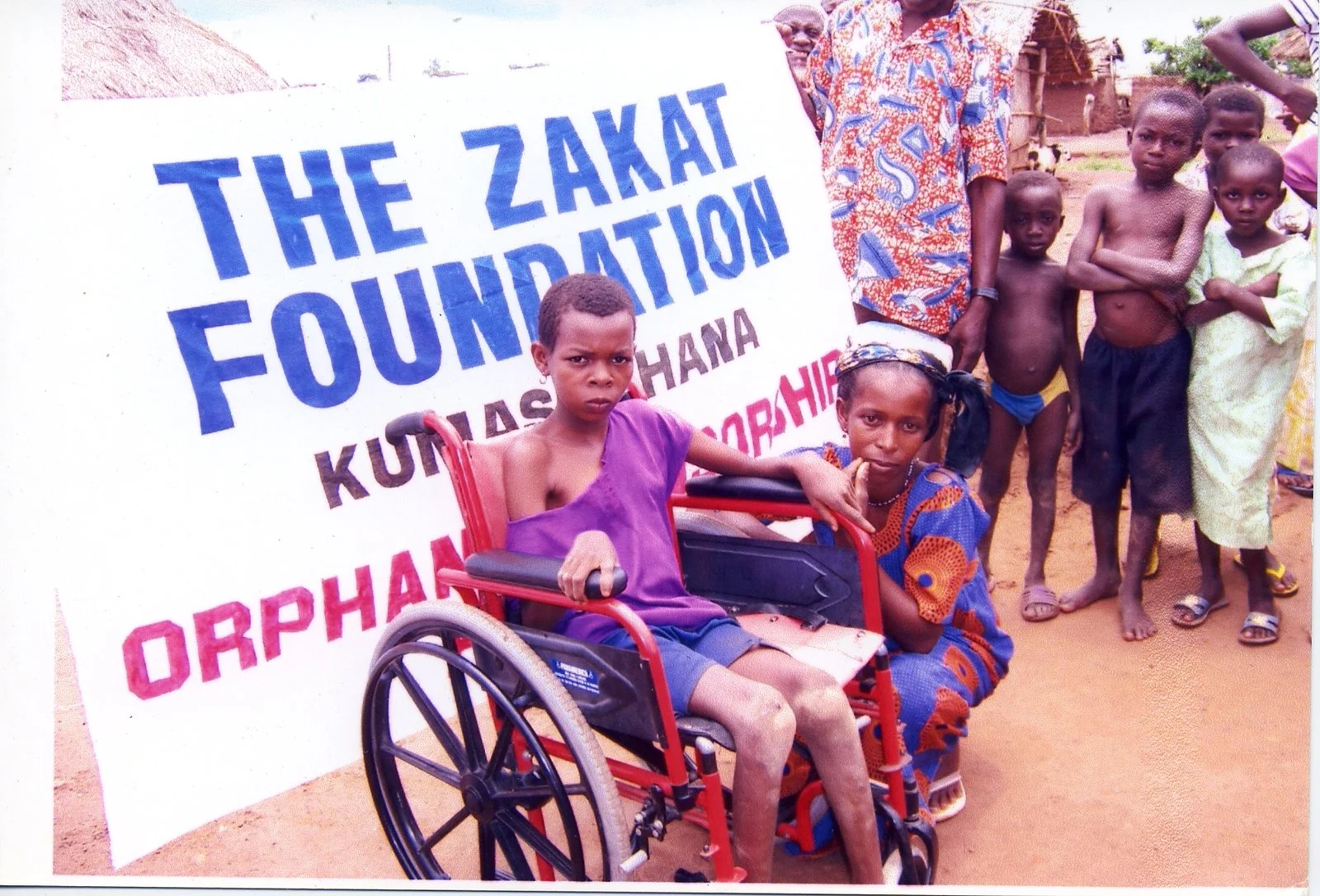 Children and adults standing outdoors next to a large sign that reads 'The Zakat Foundation' with other text underneath, some children are barefoot, and one child with a disability is in a wheelchair.