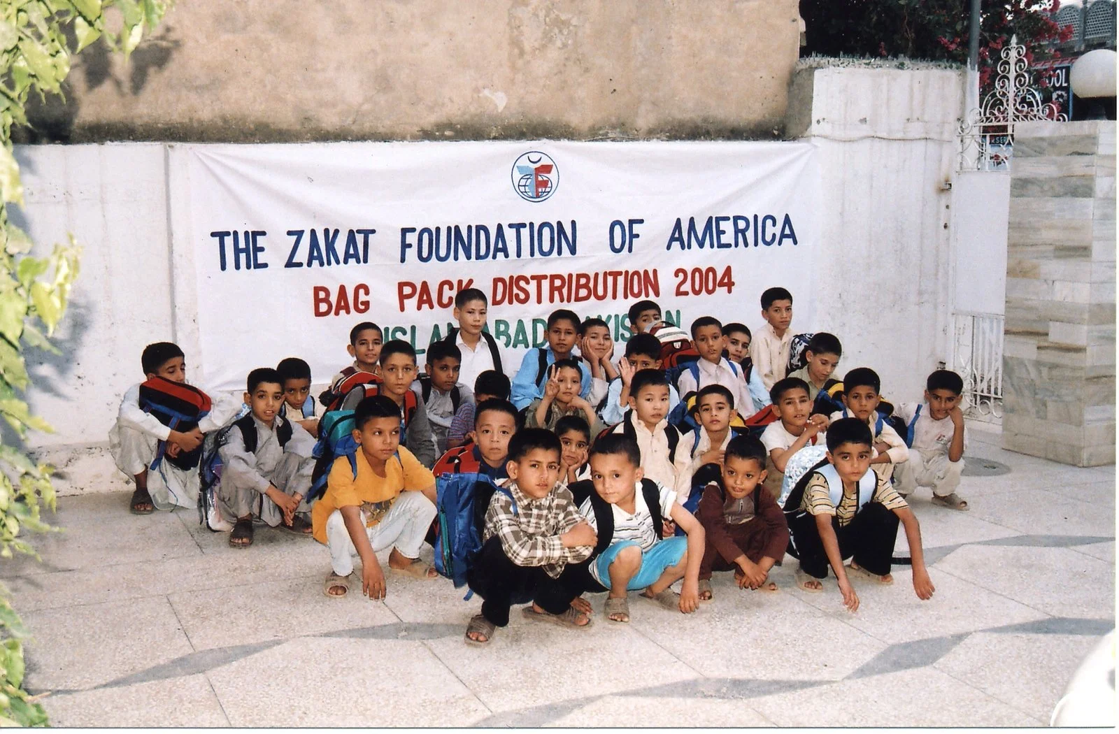 Group of young boys with backpacks sitting and standing in front of a banner with the text 'The Zakat Foundation of America Bag Pack Distribution 2004'.