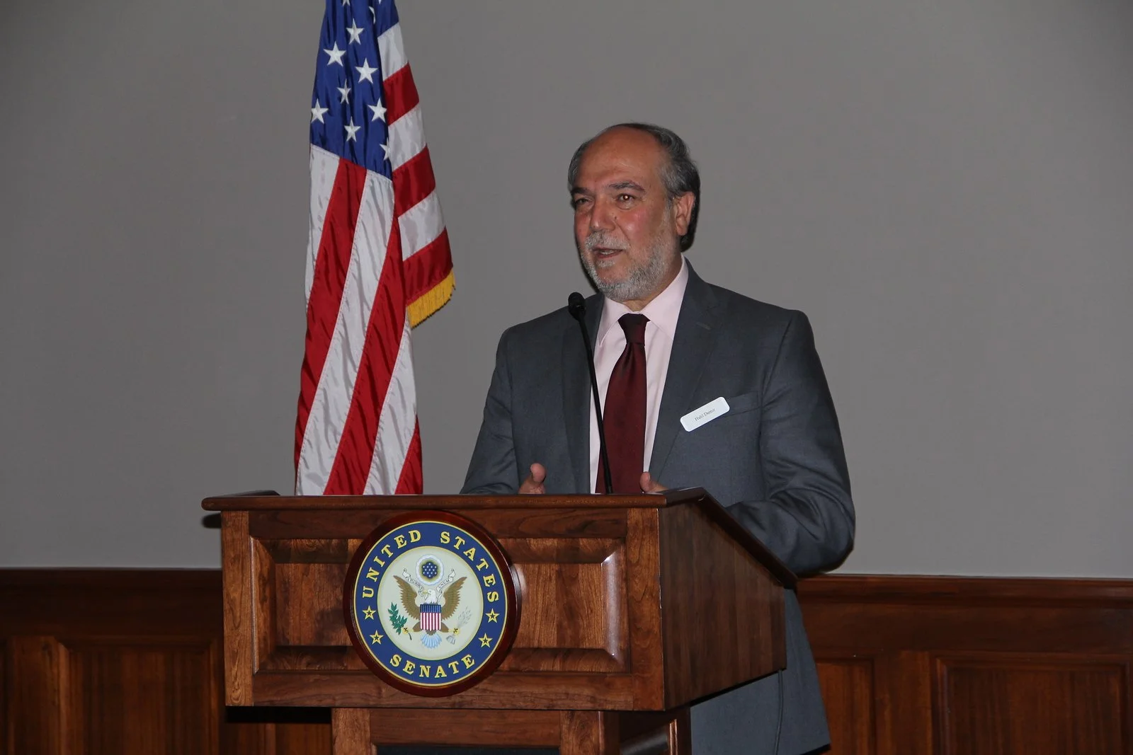 Halil Demir in a dark suit and maroon tie speaking at a wooden podium with the Seal of the United States Senate. An American flag stands behind him to the left.