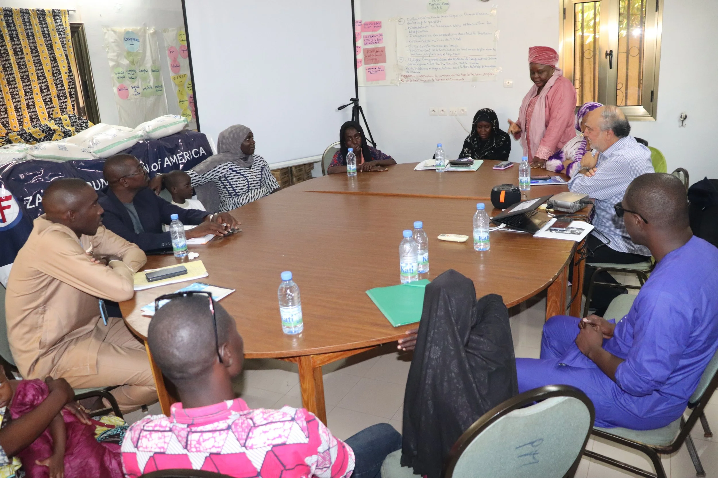 A group of people seated around a large, U-shaped conference table in a meeting room. Some participants have notebooks, water bottles, and electronic devices. One woman is standing and speaking, while others listen attentively. The room has white walls with posters, and a window lets in natural light.