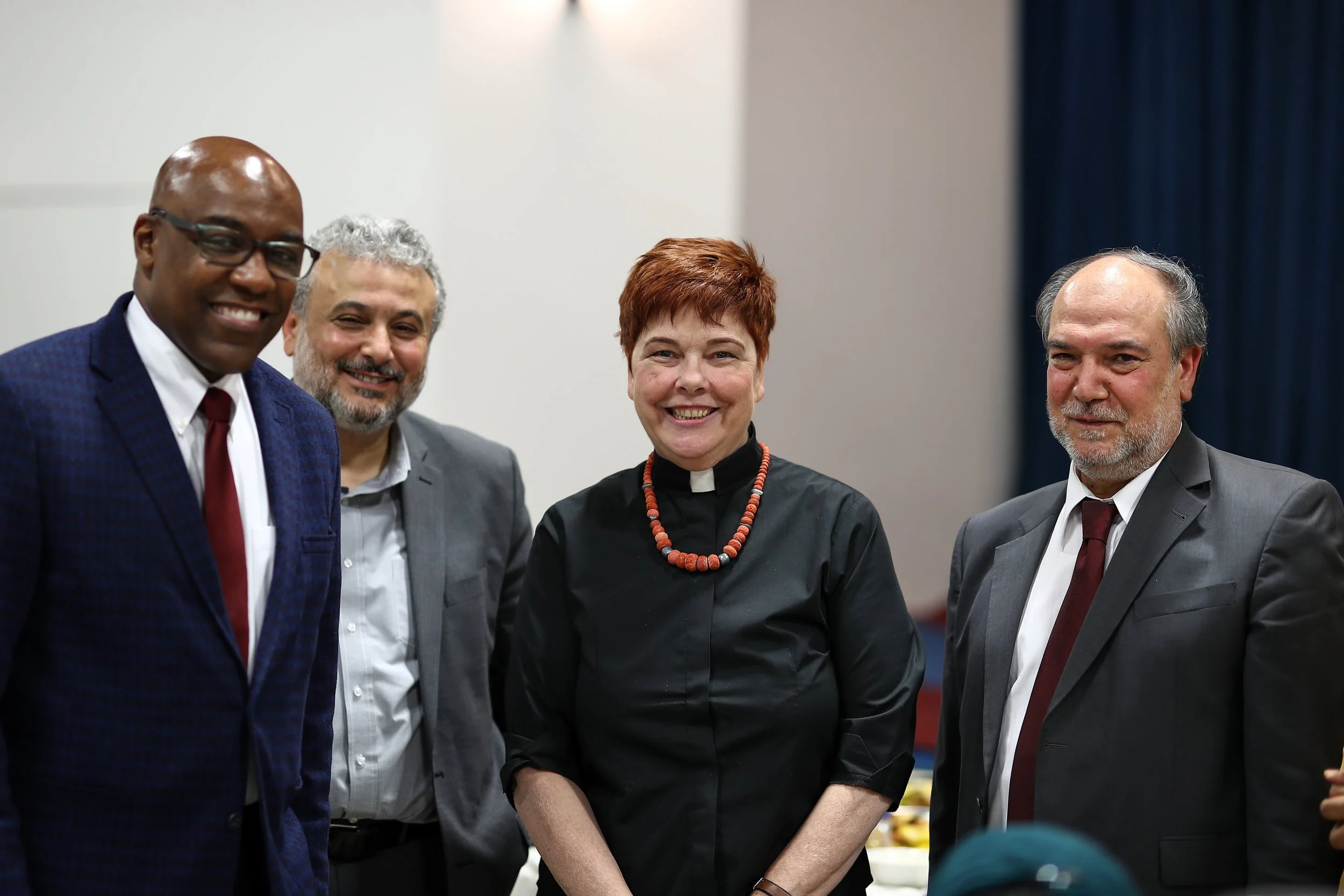 Group of four smiling people in formal attire at an indoor event, with a woman in the center wearing a black dress and a red beaded necklace.