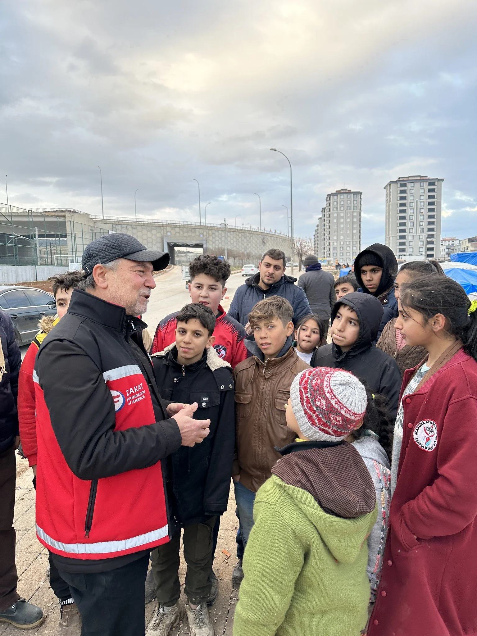 A man with a gray beard and black cap, wearing a red and black jacket, talking to a group of children and teenagers outdoors on a cloudy day. The background shows tall buildings and a road.
