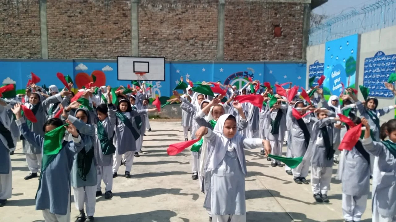 Group of school children performing a dance or activity outdoors while holding red and green cloths, wearing uniforms and headscarves, with a mural and brick wall in the background.