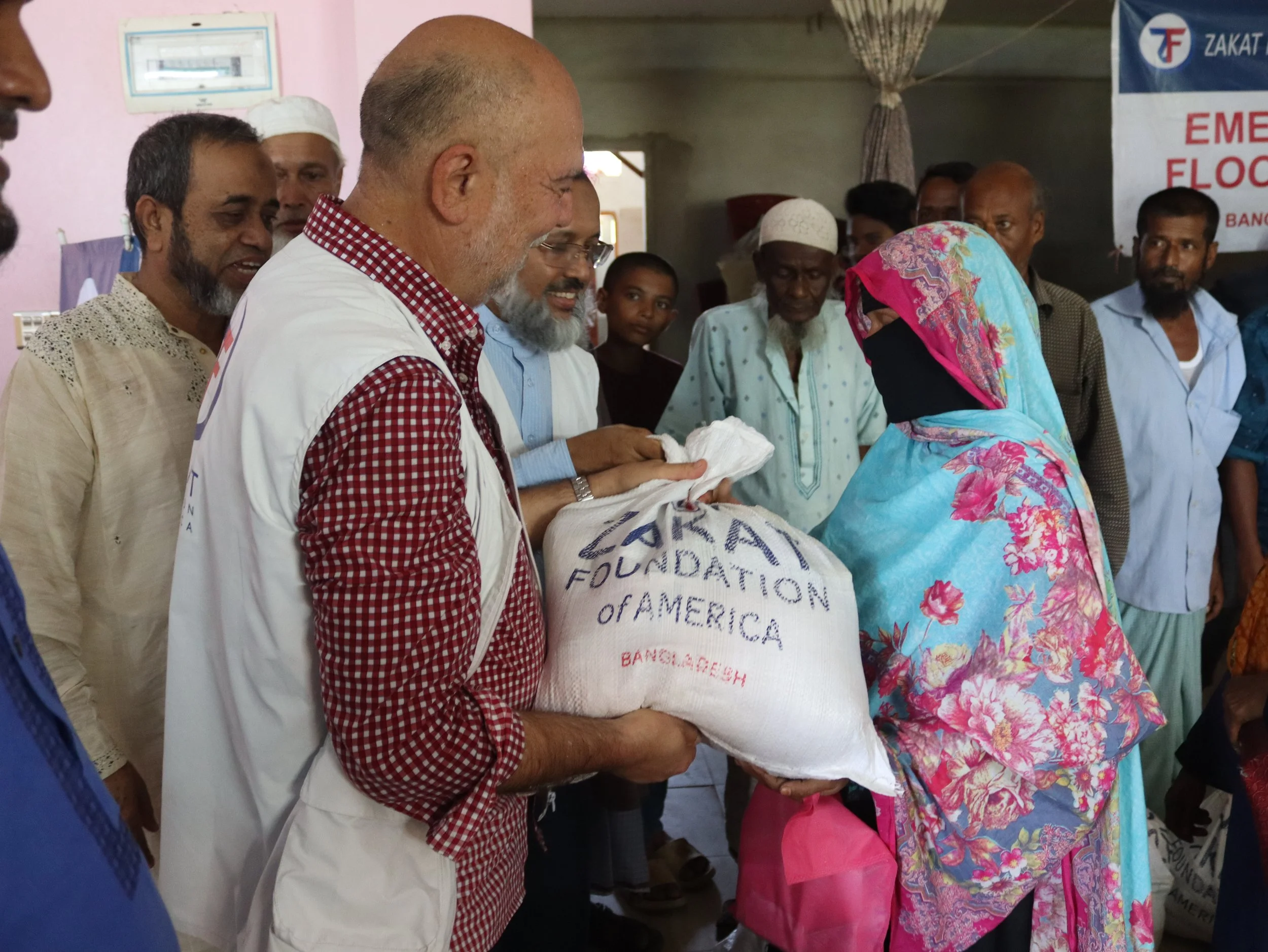 Halil Demir from Zakat Foundation of America handing a bag of aid to a woman in a floral headscarf, surrounded by men in traditional clothing in a humanitarian aid setting.