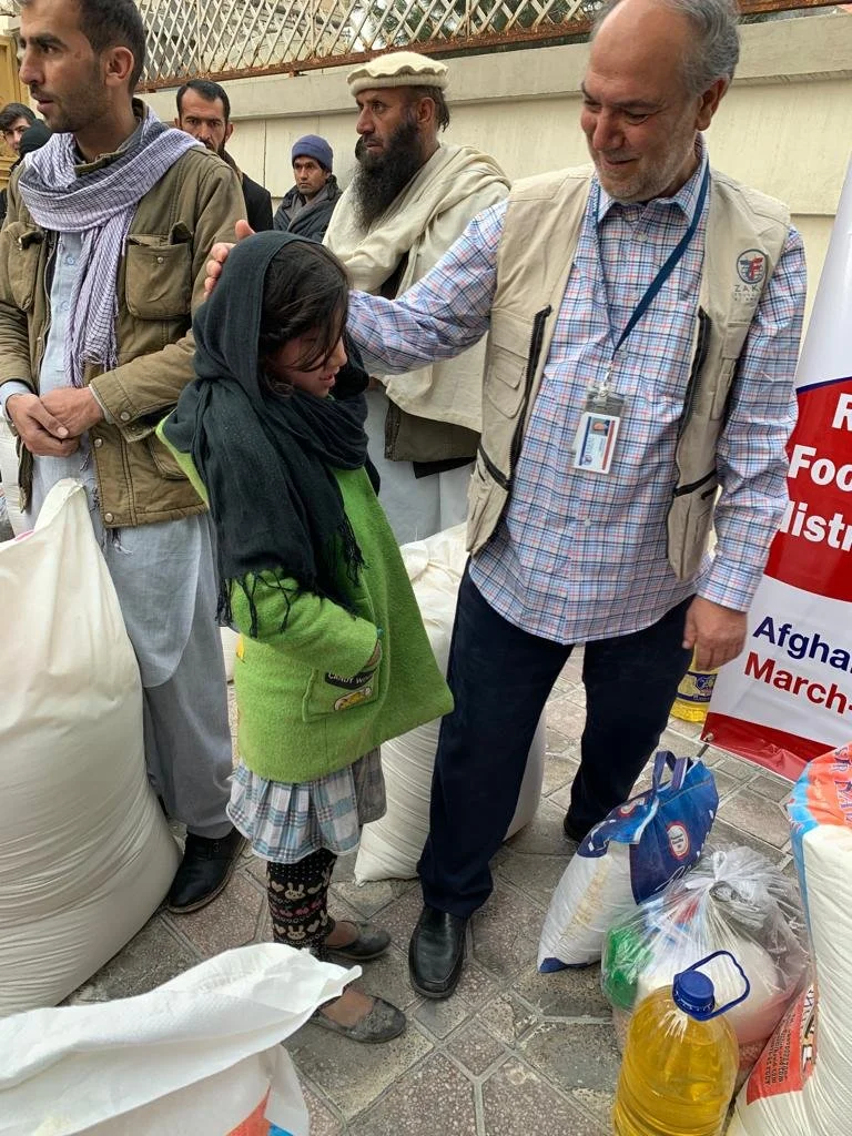 A man in a vest and plaid shirt, with a lanyard around his neck, gently patting a young girl on the head during a humanitarian aid event. The girl wears a black headscarf, a green jacket, and a plaid skirt. Several other people stand nearby, and large bags of supplies are visible on the ground around them.